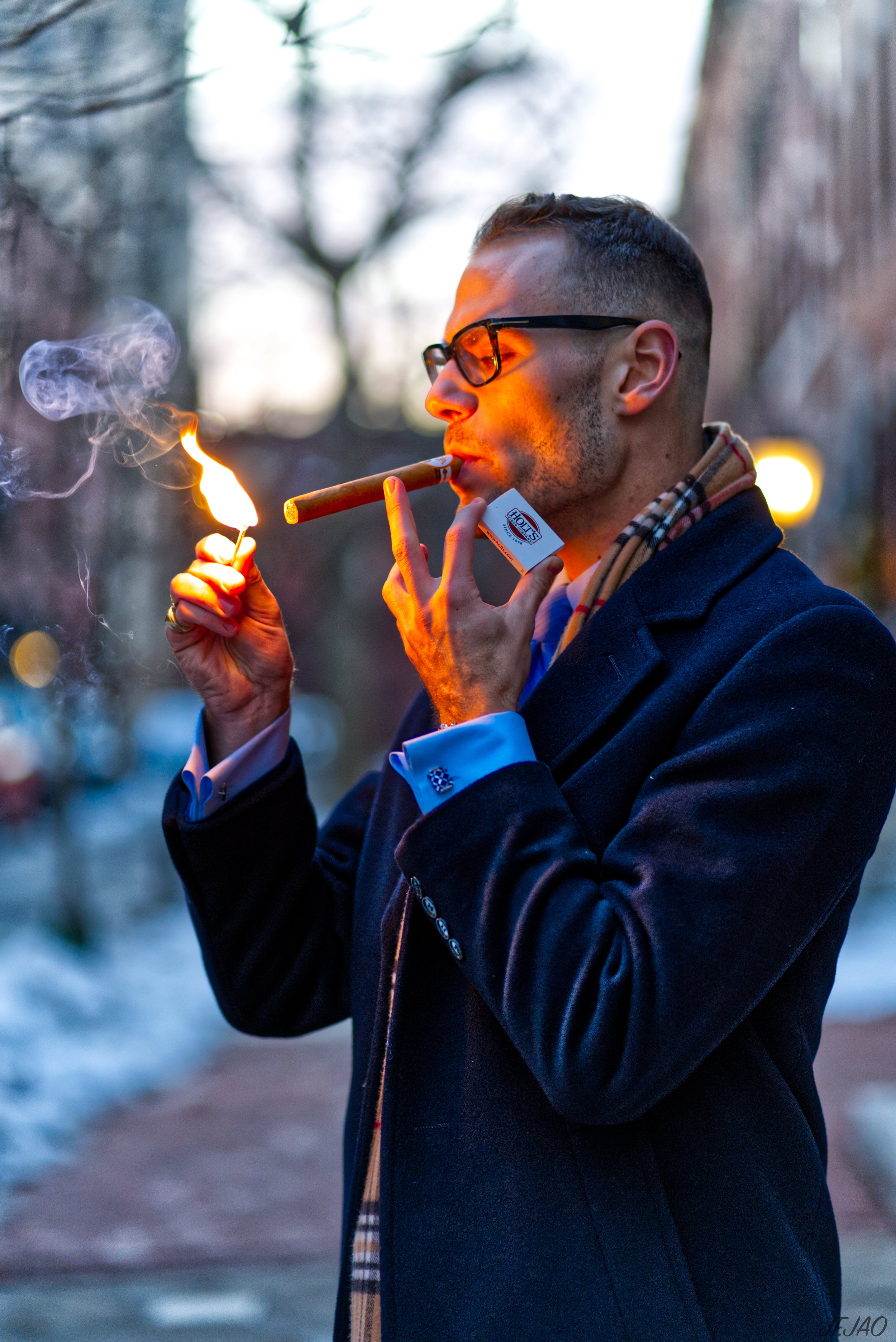 Man lighting a cigar outdoors with a match, wearing a coat and scarf, glasses, and a ring.