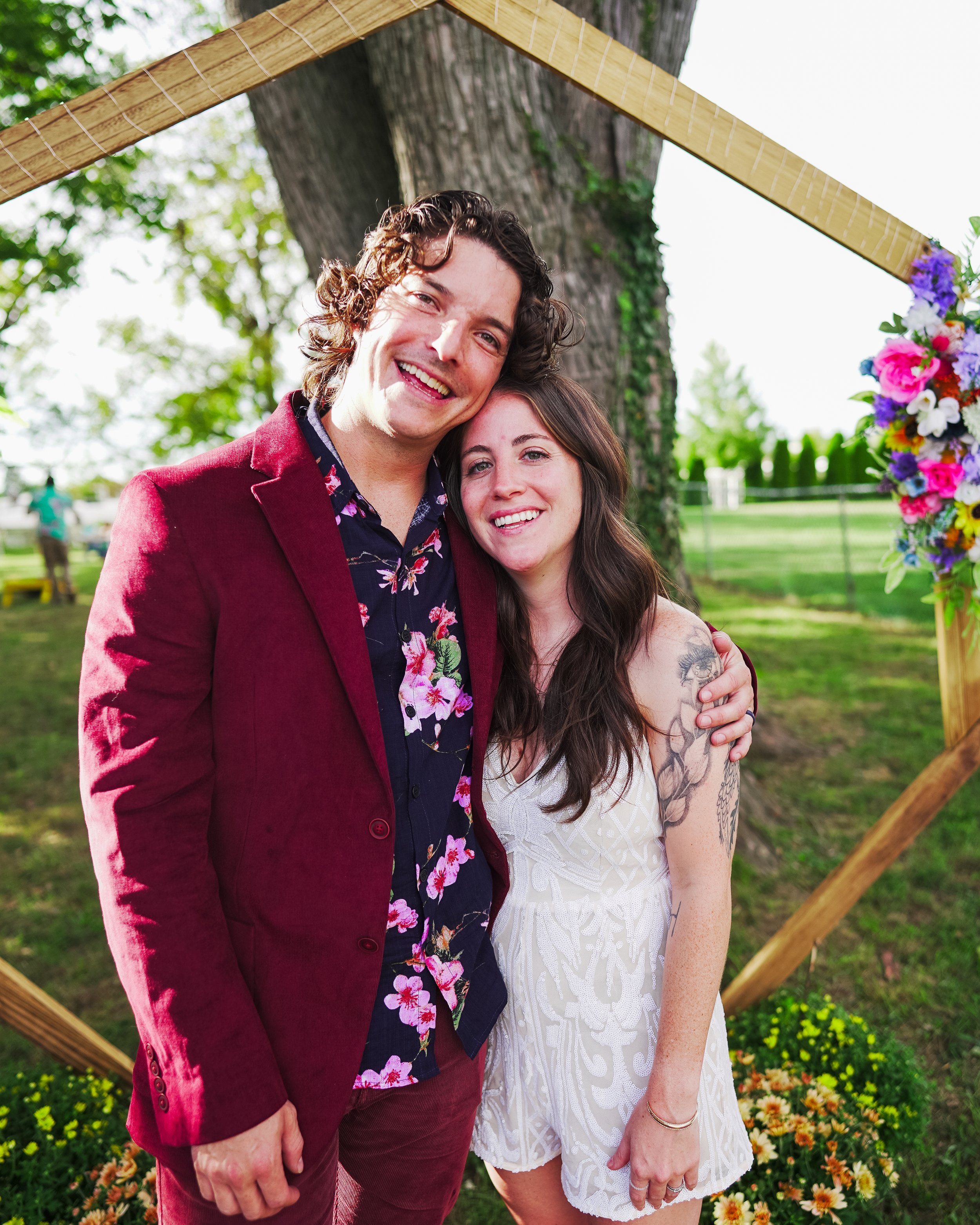 A smiling couple posing outdoors in front of a large tree, framed by a wooden structure decorated with colorful flowers. The man is wearing a maroon suit jacket and floral shirt, while the woman is wearing a white lace dress with tattoos on her arm.