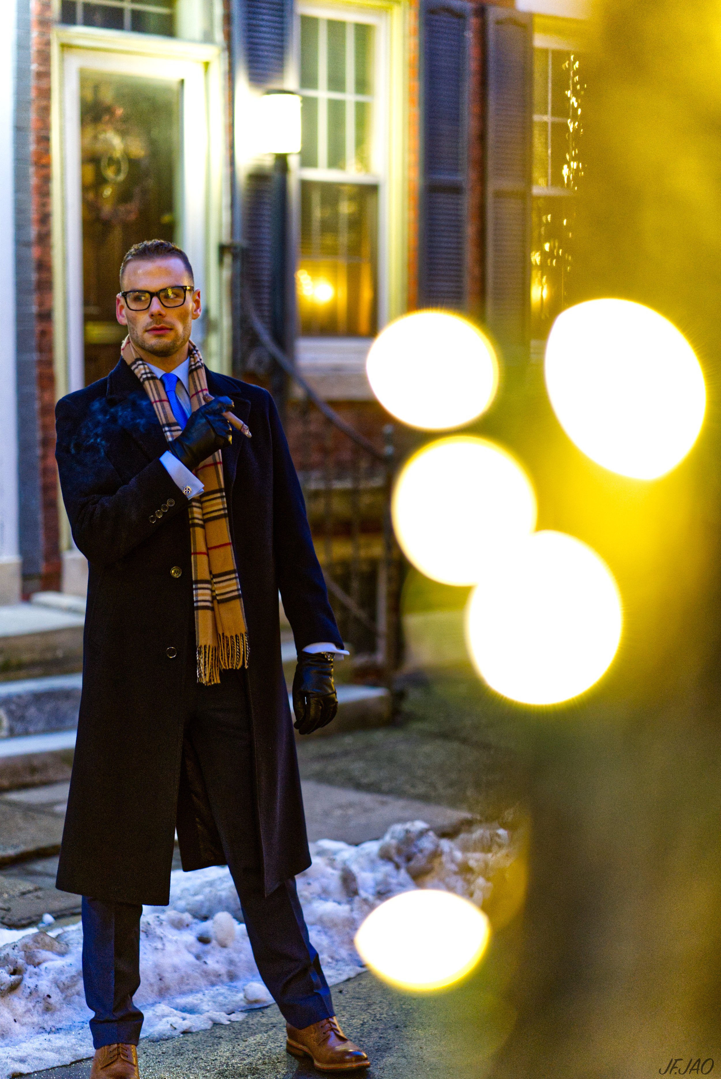 Man in a coat and scarf standing outdoors by a building with lit windows, holding a cigar, with blurred yellow lights in the foreground and snow on the ground.