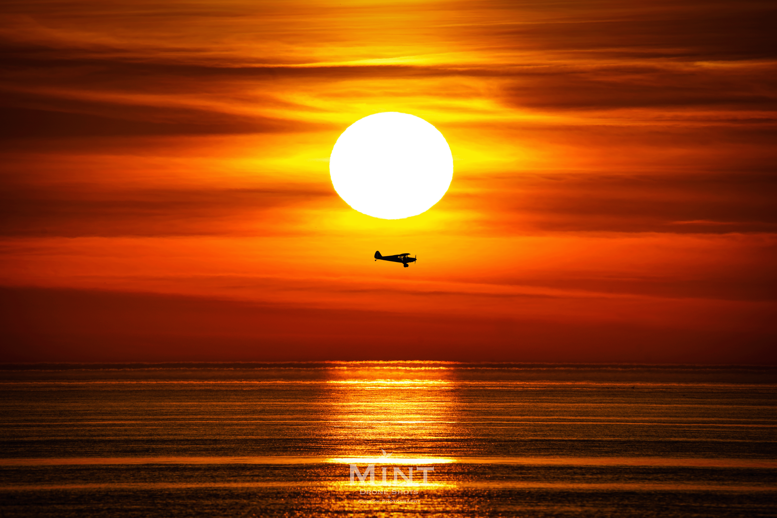Airplane silhouette flying in front of setting sun over ocean with orange sky.