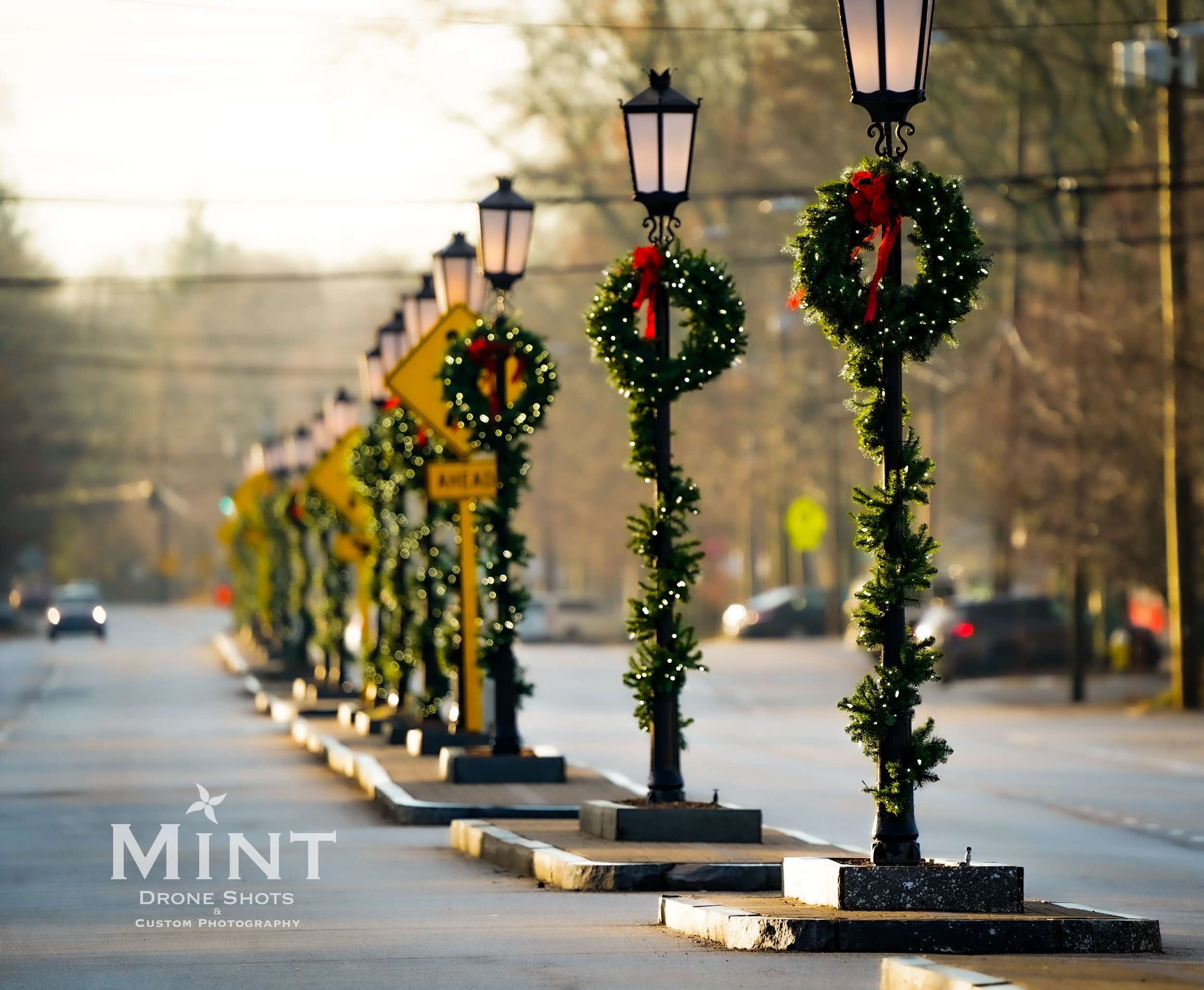 Street lined with decorative lampposts wrapped in garland and wreaths, featuring red bows and lights, during a holiday season.