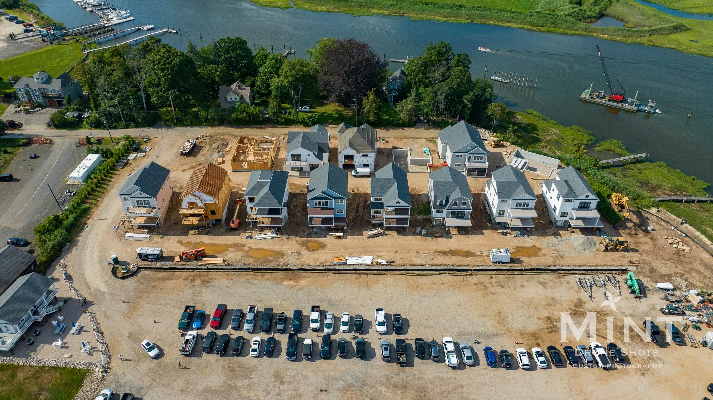 Aerial view of a housing development near a river with several under-construction houses, parked vehicles, and surrounding greenery.