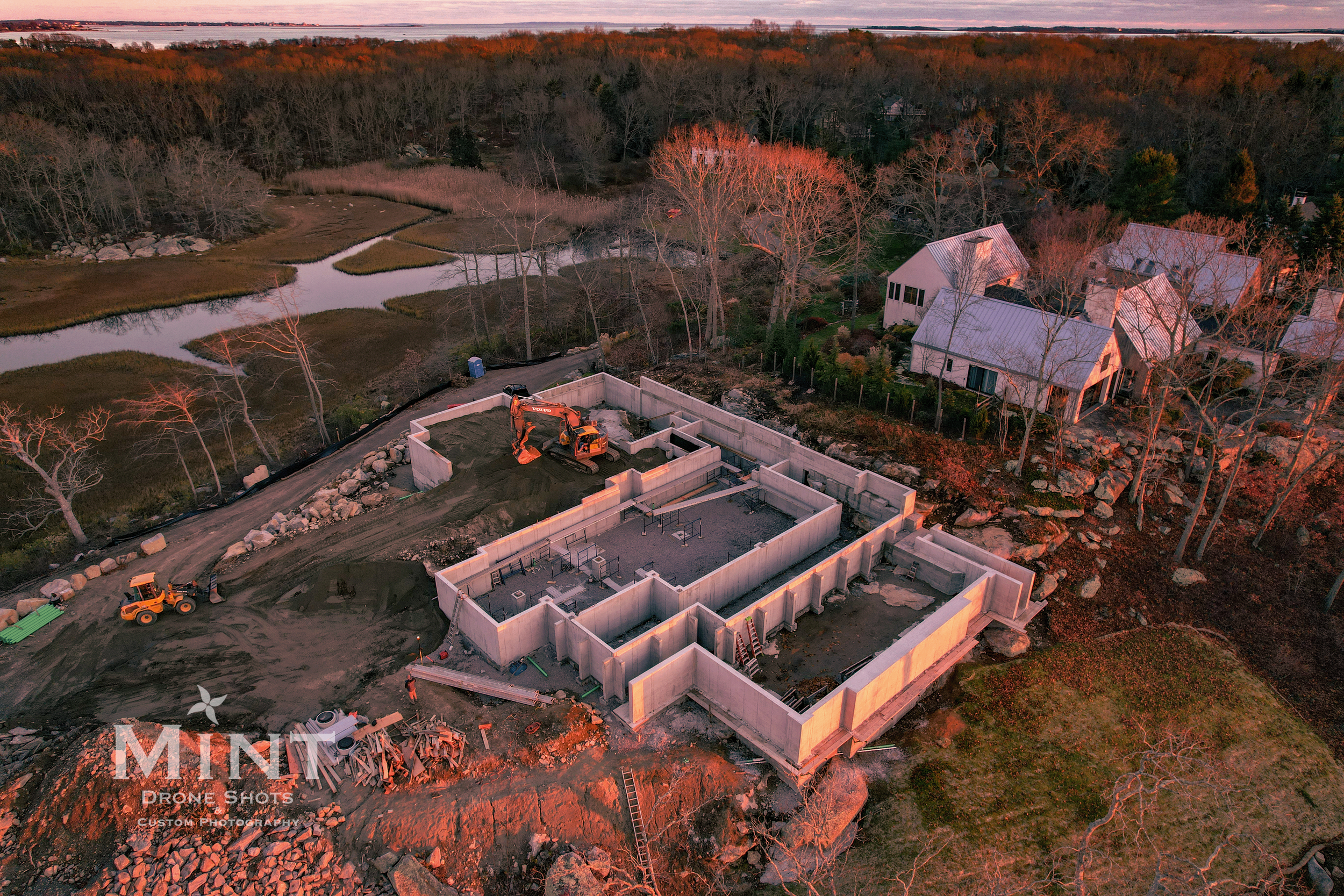 Aerial view of a construction site with concrete foundation, excavators, and nearby houses during sunset or sunrise.