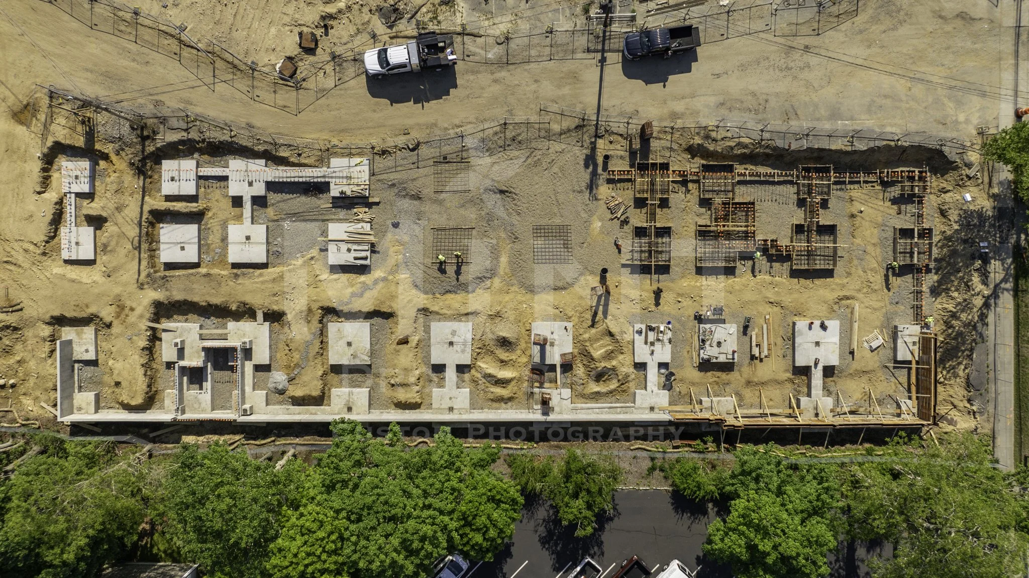 Aerial view of a construction site with concrete foundations and structures, surrounded by trees and vehicles parked along a road.