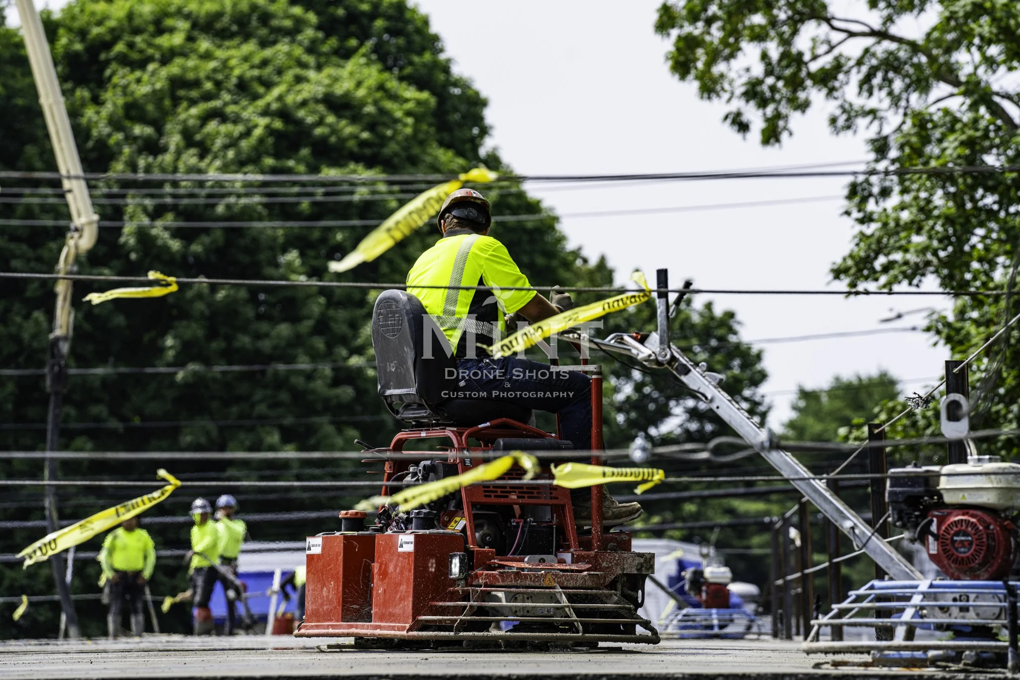 Construction worker operating a trowel machine on a concrete surface, surrounded by caution tape and additional workers in the background. Trees and utility lines are visible.