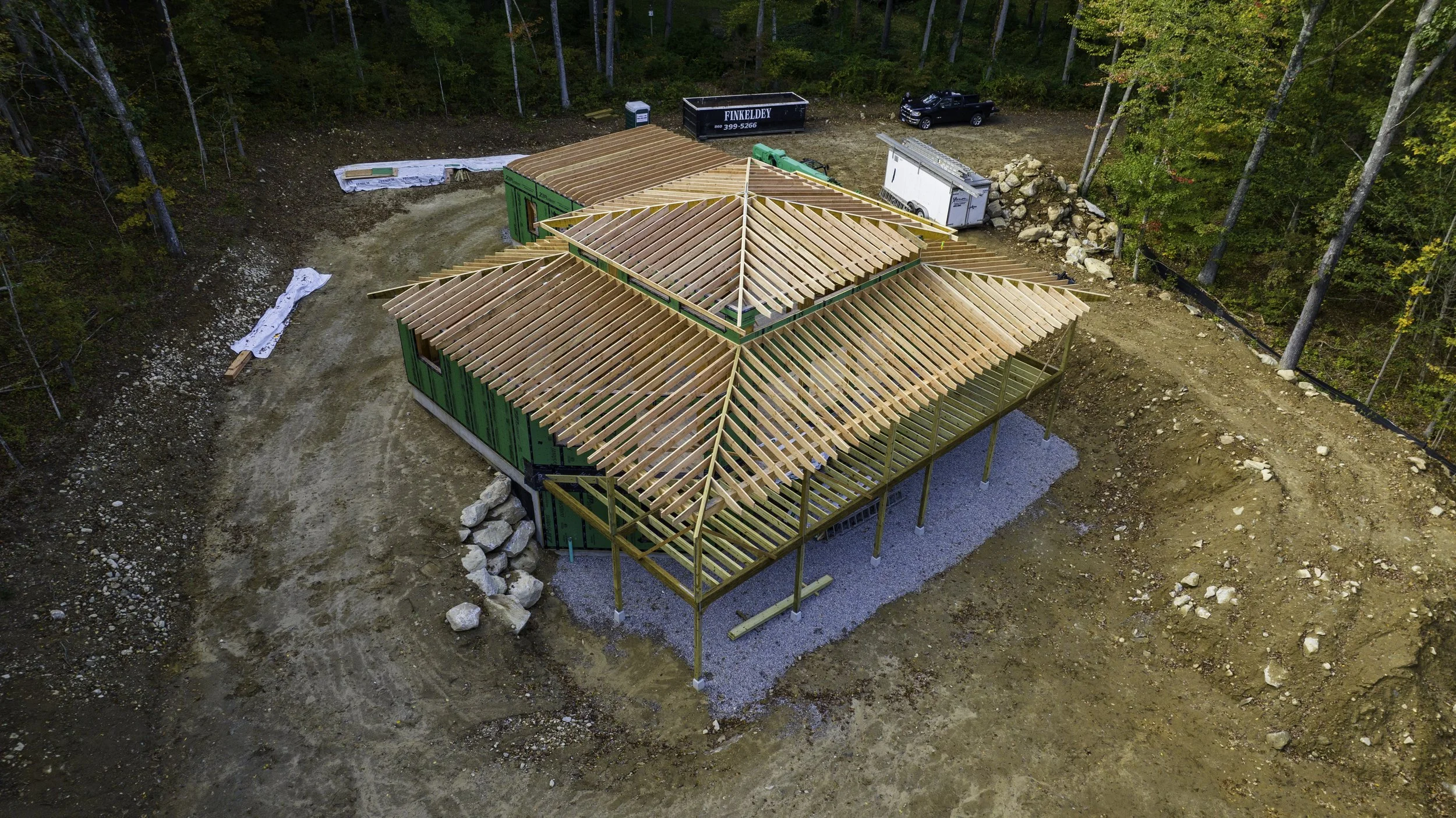 Aerial view of an unfinished wooden house under construction in a forested area, featuring exposed roof beams and surrounding open space.