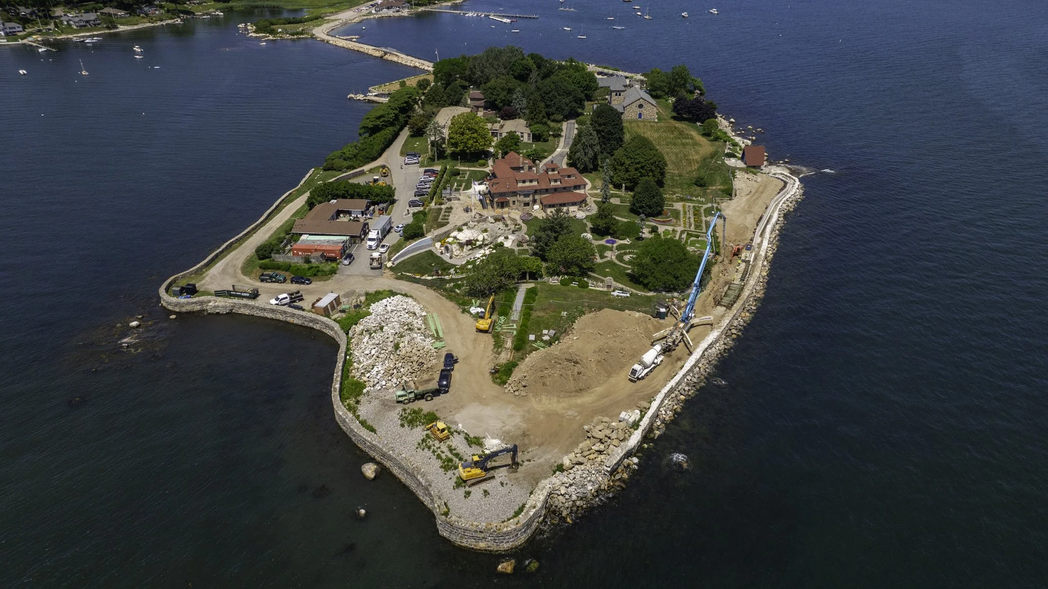 Aerial view of a small island with residential buildings, gardens, and construction equipment near the shore. The island is surrounded by water, and vehicles are parked along a road. There is visible construction activity with machinery and piles of 