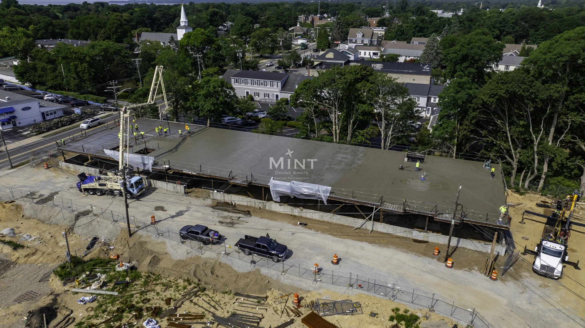 Aerial view of a construction site with workers smoothing a large concrete slab. A concrete pump truck and construction equipment are visible. There are trees and buildings in the background.