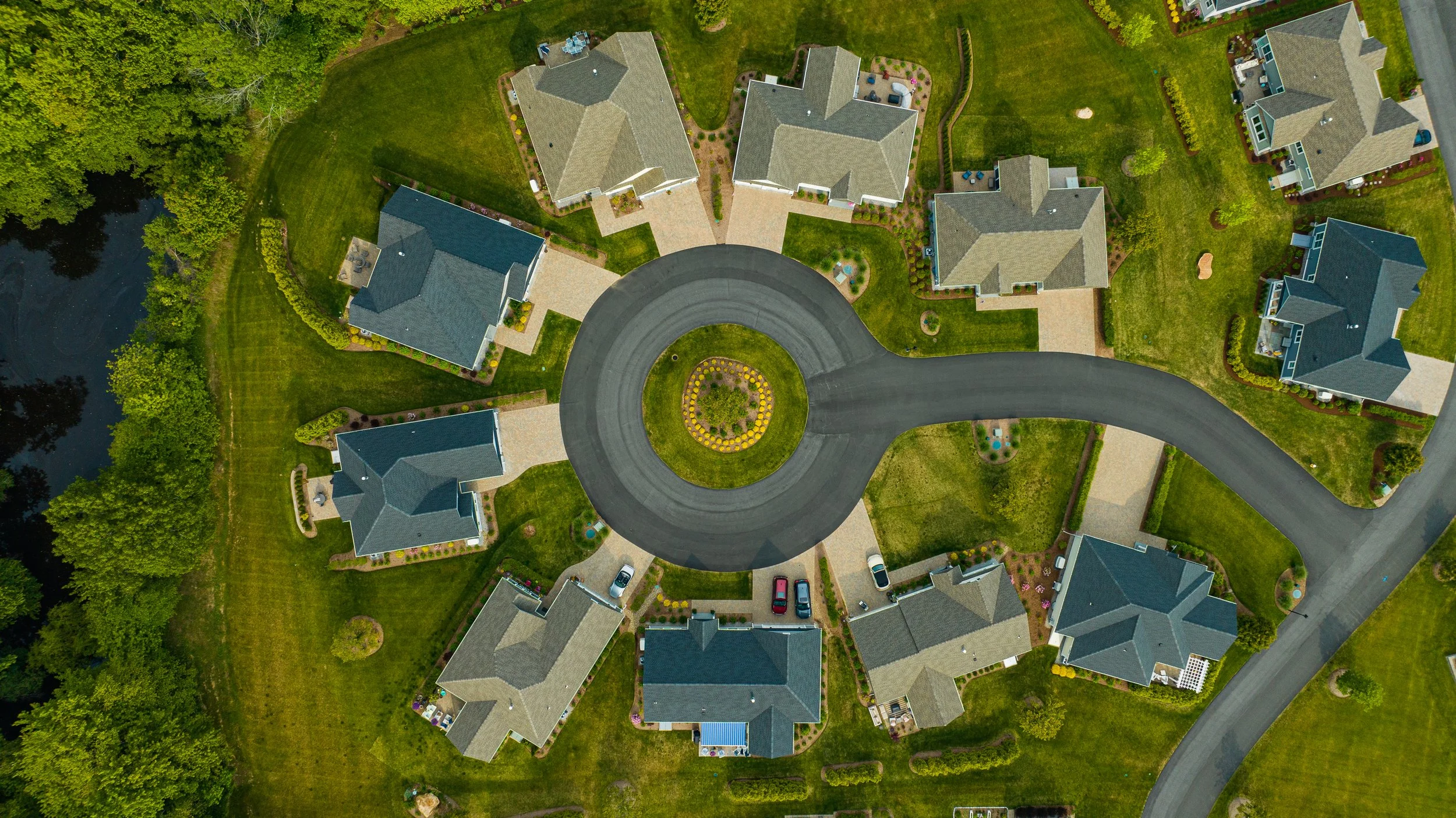 Aerial view of a suburban cul-de-sac with houses surrounding a circular road. The homes are evenly spaced with well-maintained lawns, driveways, and landscaping. There is a small grassy area with trees outside the circle.