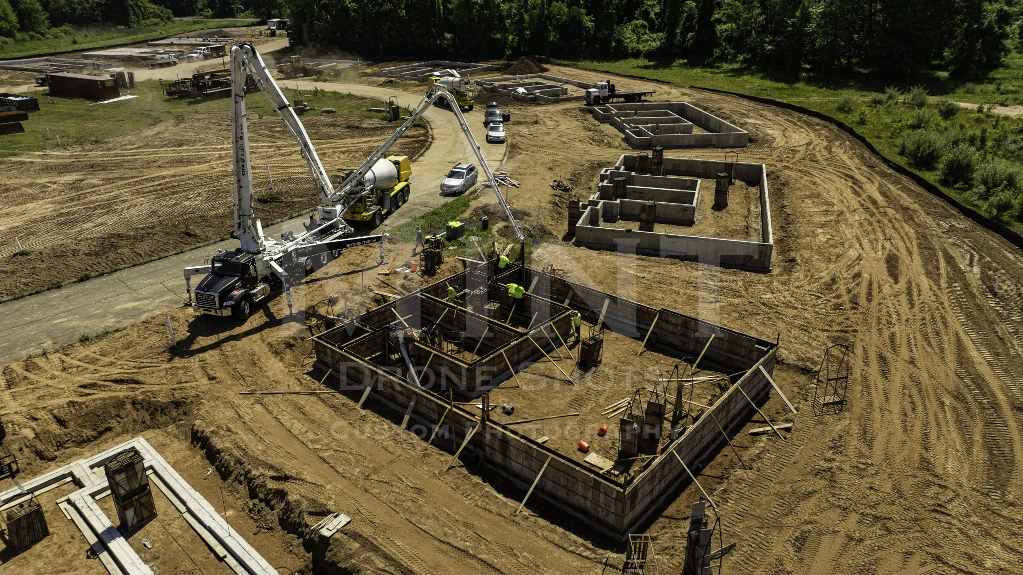 Aerial view of a construction site with workers, concrete forms, and heavy machinery, including a concrete pump truck, surrounded by dirt and greenery.