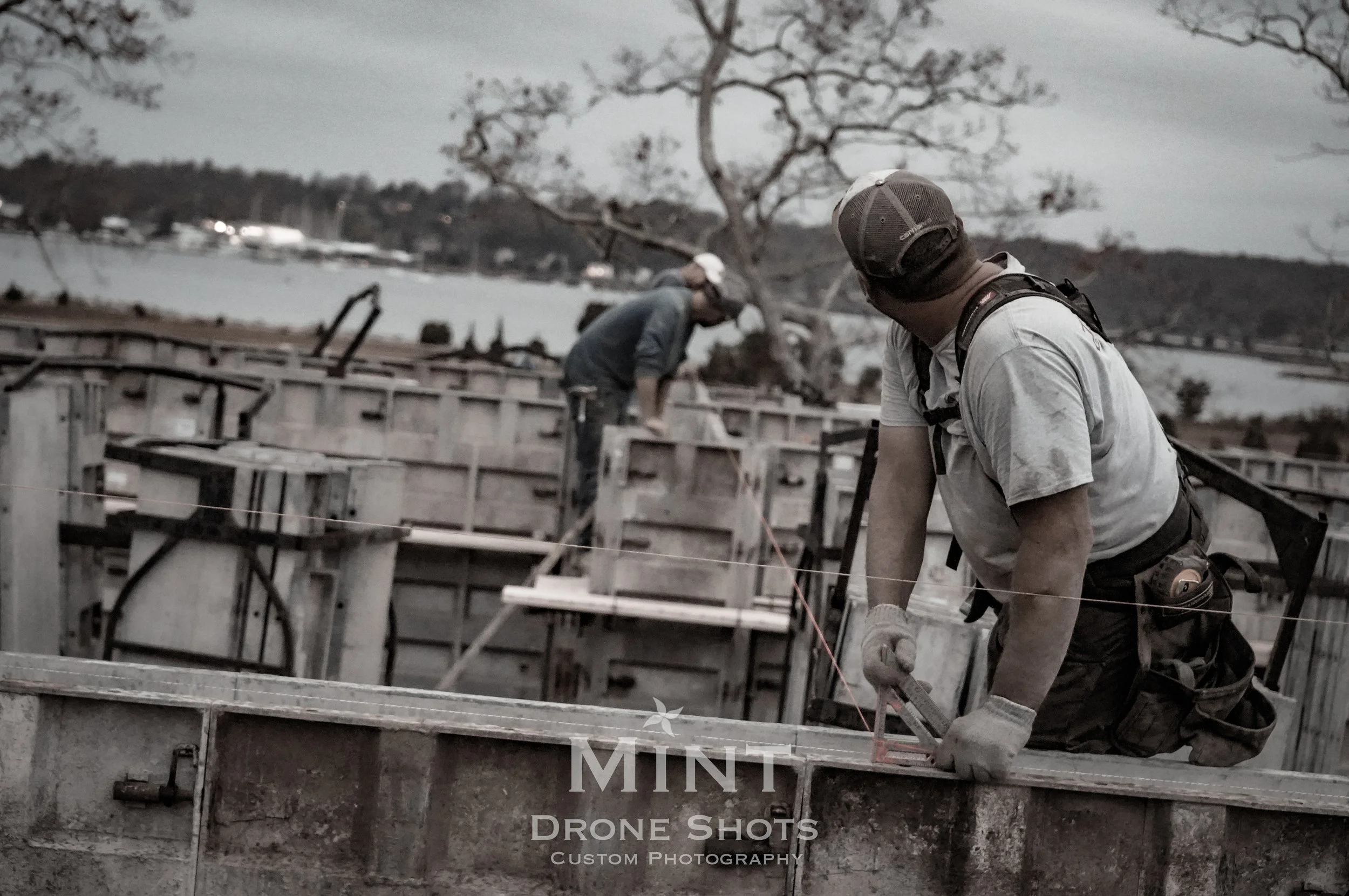 Two construction workers wearing caps and work gear performing tasks on a building site near a body of water, with trees and a cloudy sky in the background.