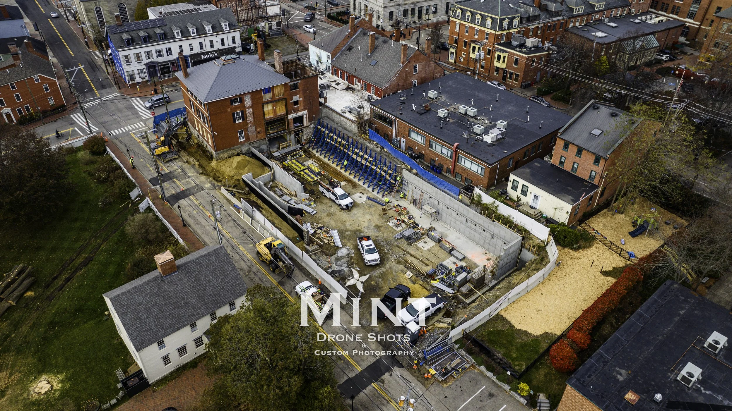Aerial view of a construction site with surrounding brick buildings, streets, and vehicles. Workers and construction equipment are visible within the fenced-off area. Nearby are older residential and commercial buildings.