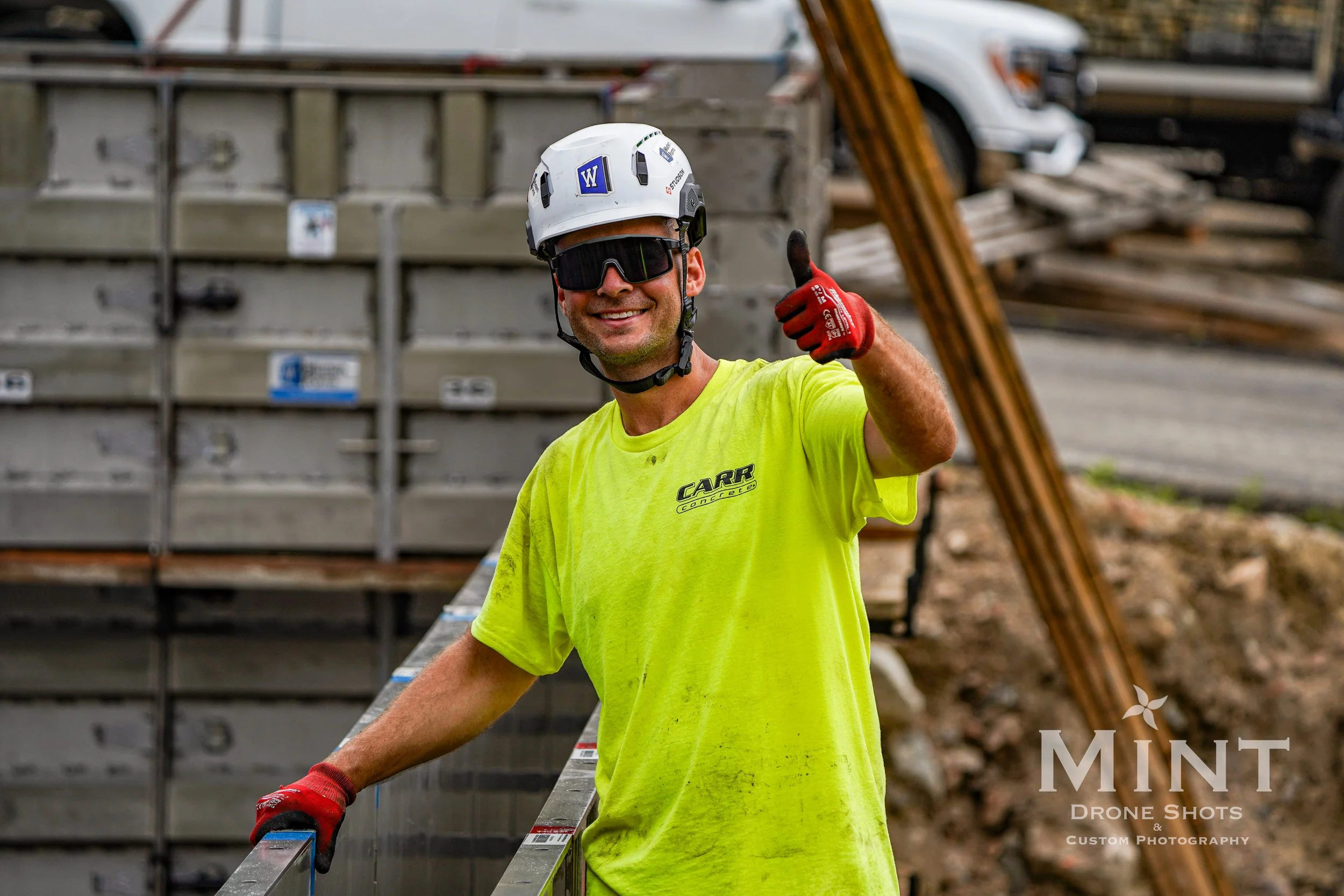 Construction worker in a yellow shirt and hard hat giving a thumbs up at a construction site.