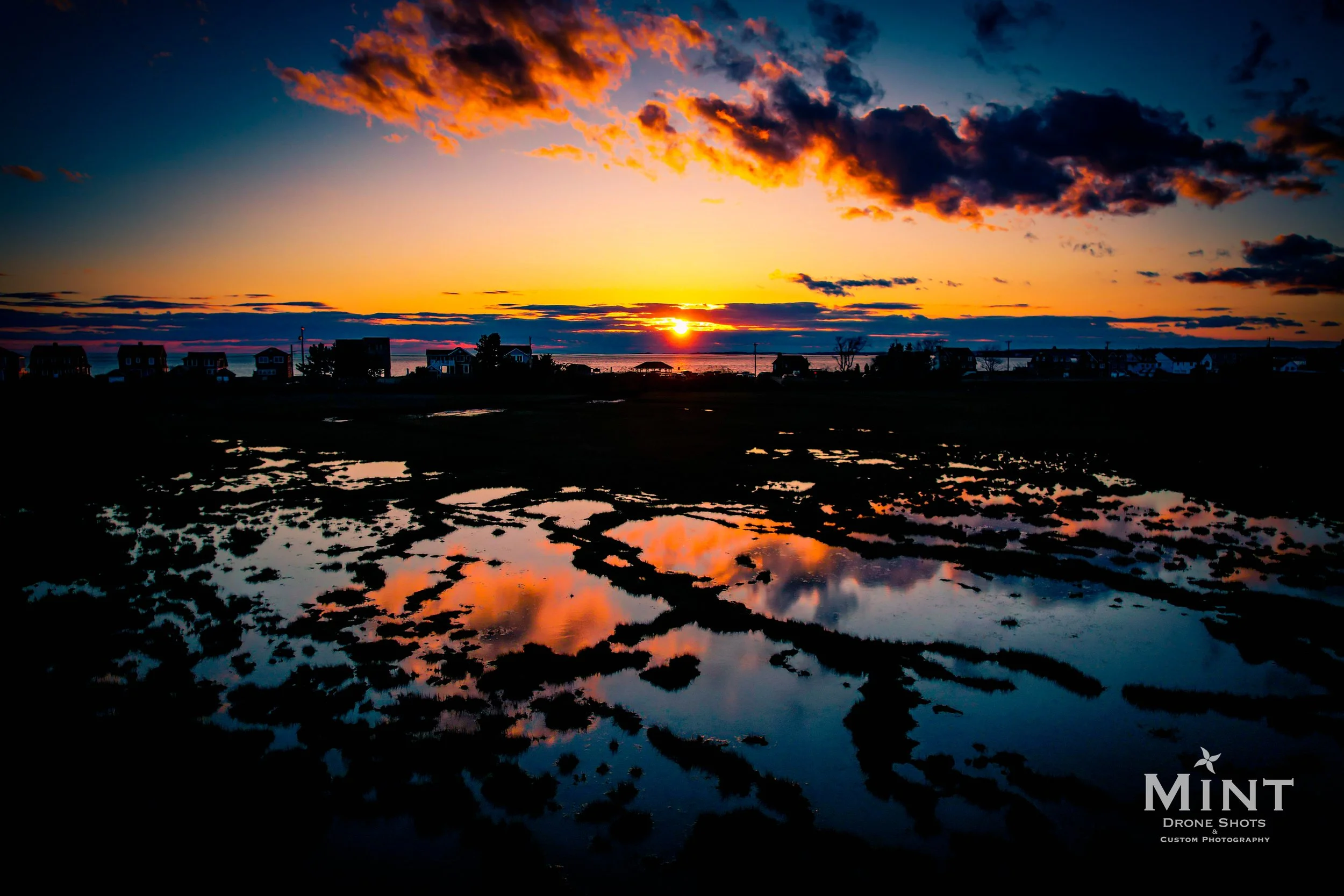 Sunset over a marsh with vibrant orange and blue sky reflections in water, silhouetted buildings in the distance.