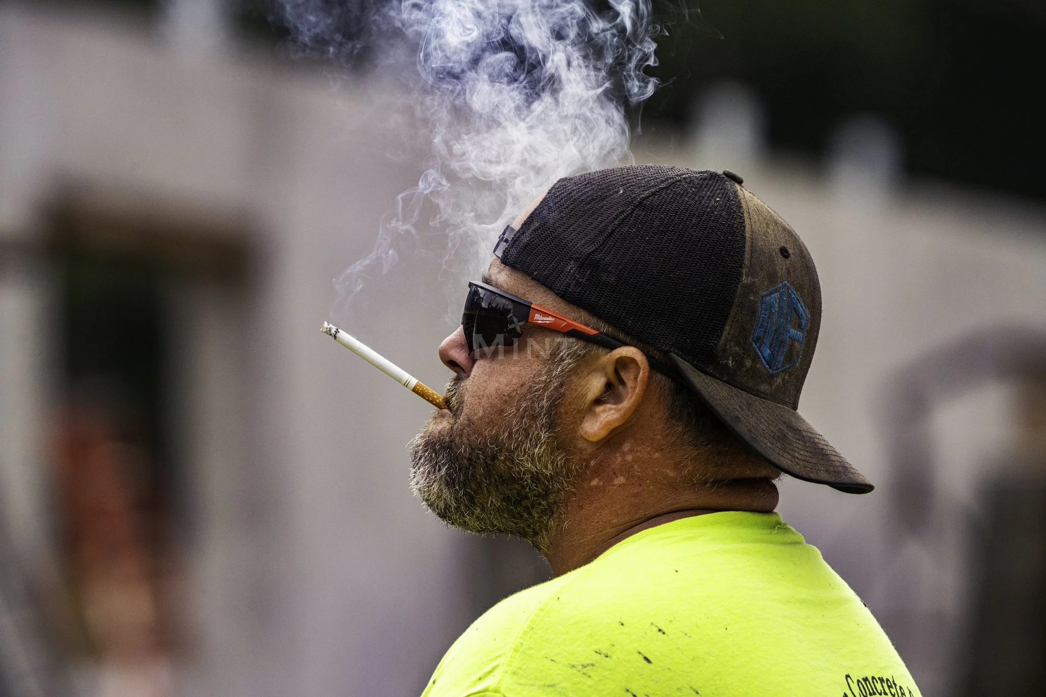 Man wearing sunglasses and a cap, smoking a cigarette, with smoke rising in the air.