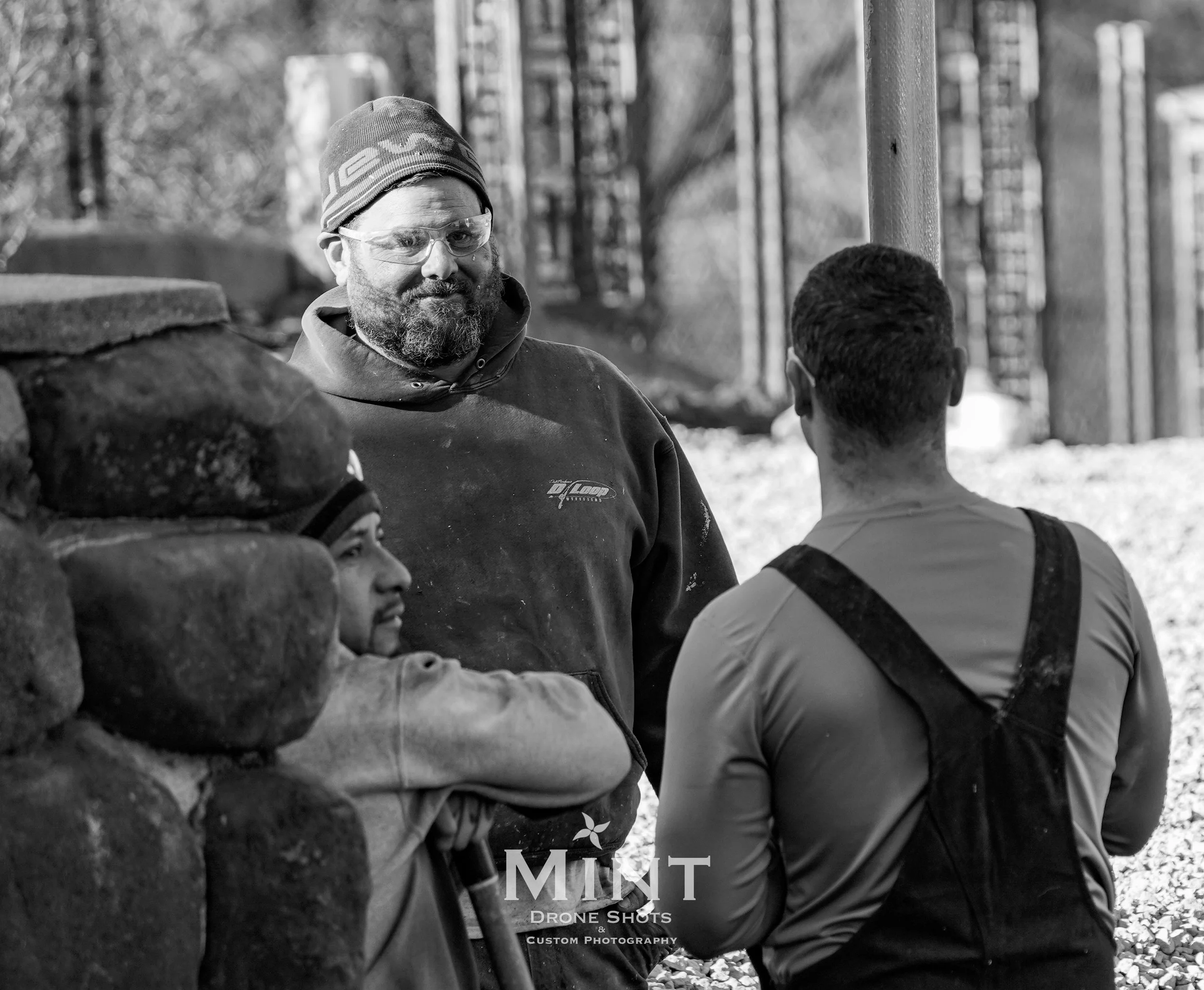 Three men in work clothes having a discussion outdoors near a stone structure.