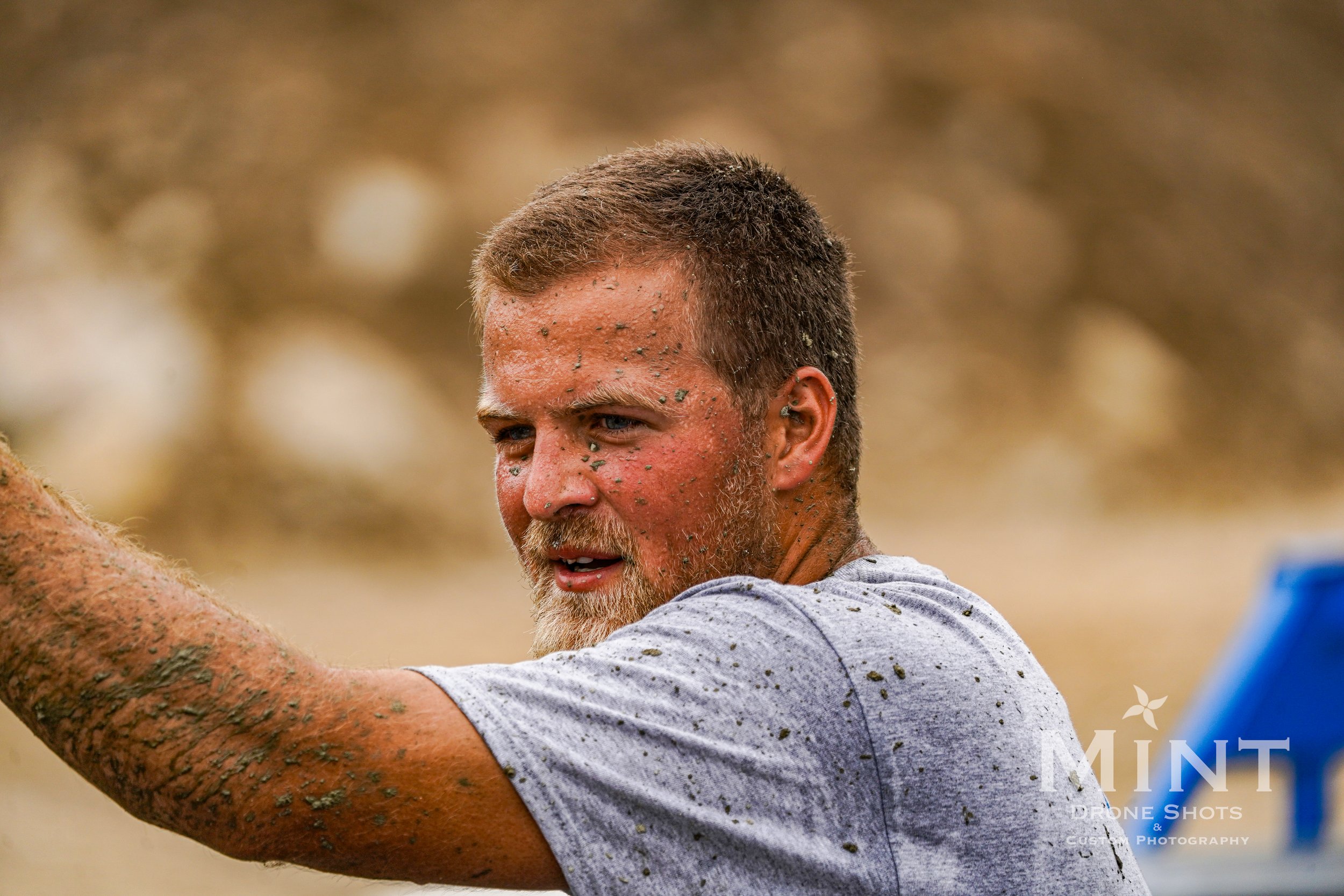 A person with short hair and a beard, wearing a gray T-shirt, covered in mud, with a focused expression, possibly participating in an outdoor event or activity.