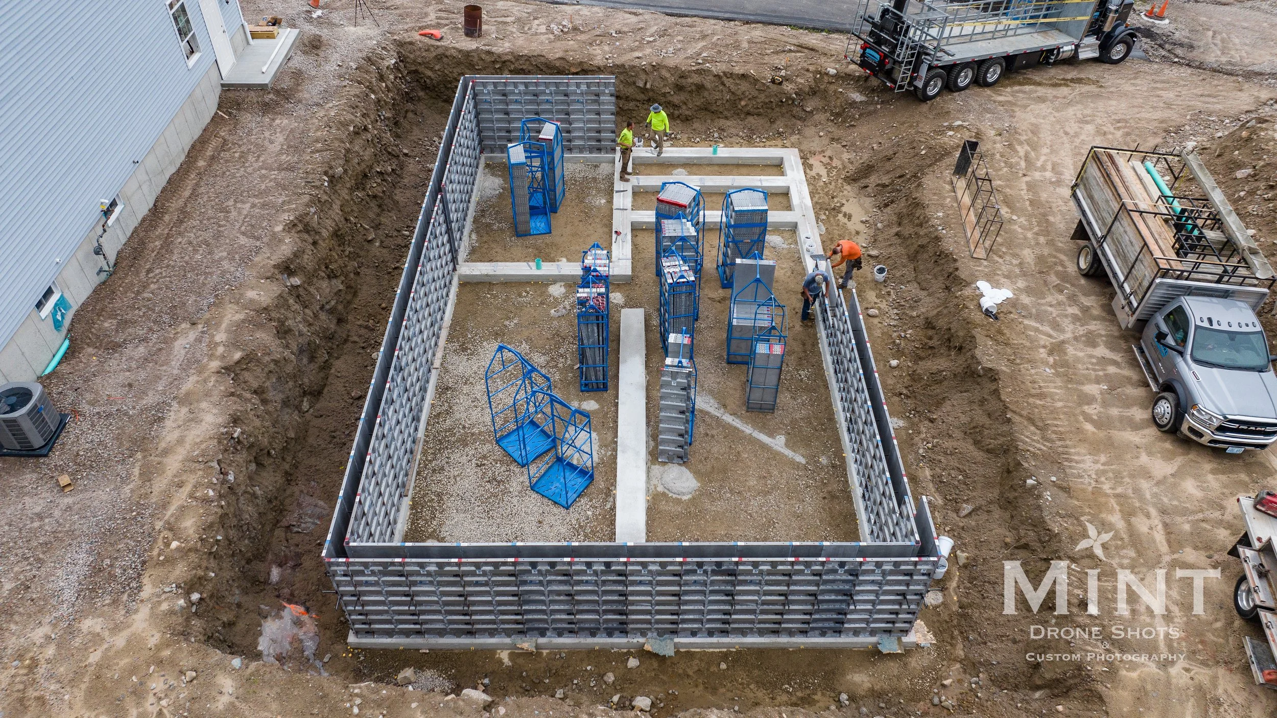 Aerial view of a construction site with foundation walls, blue scaffolding, and workers. Trucks and construction materials surround the area.
