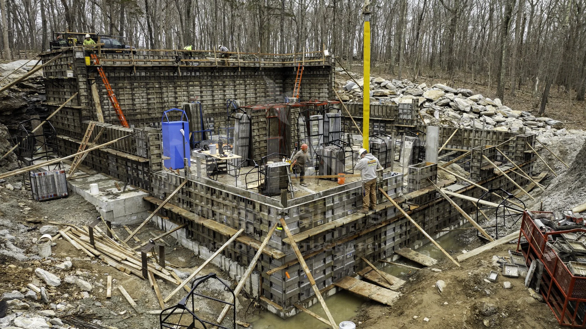 Construction site with concrete foundation and workers, surrounded by forest.