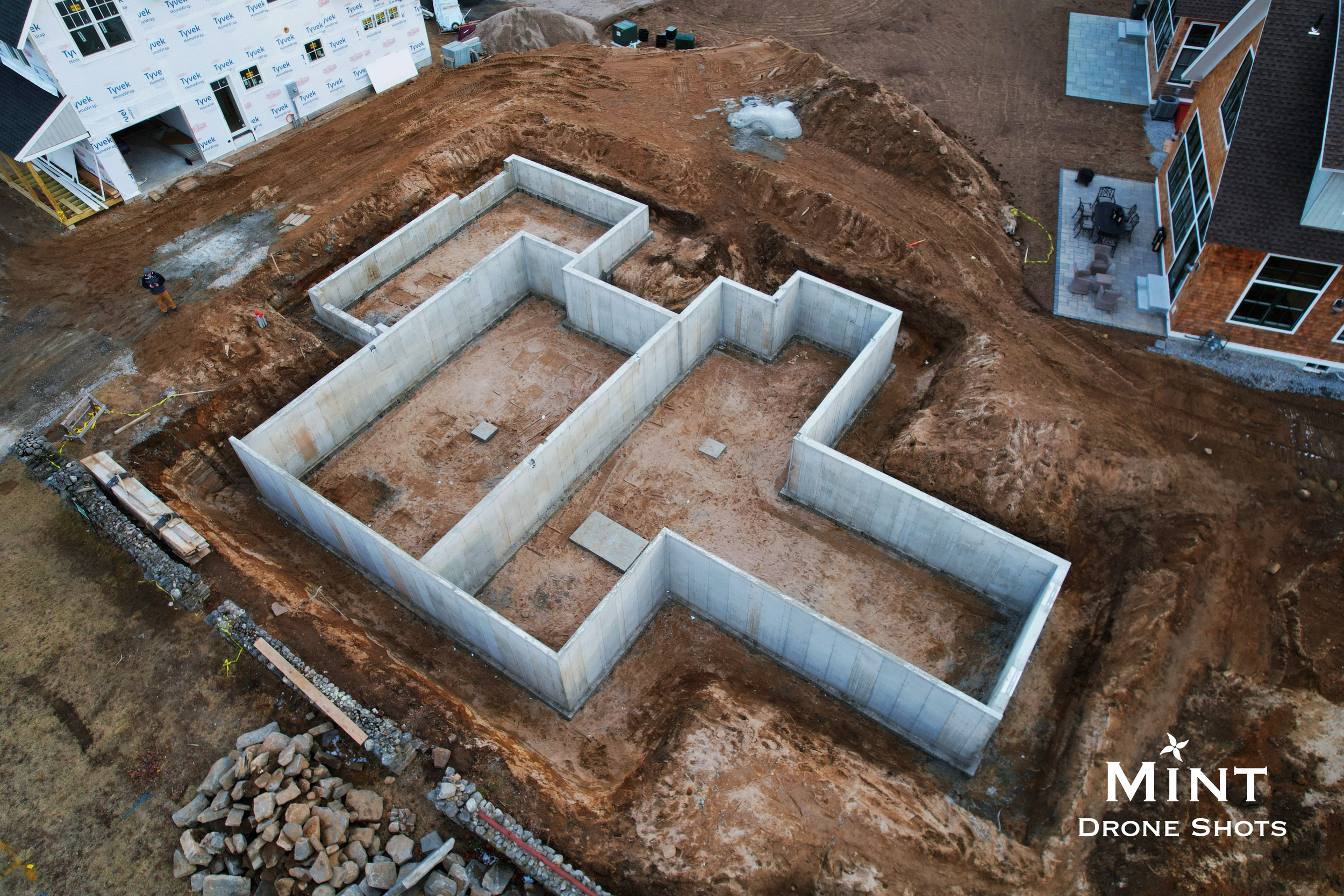 Aerial view of a building construction site with concrete foundation walls, surrounded by dirt and adjacent completed structures.