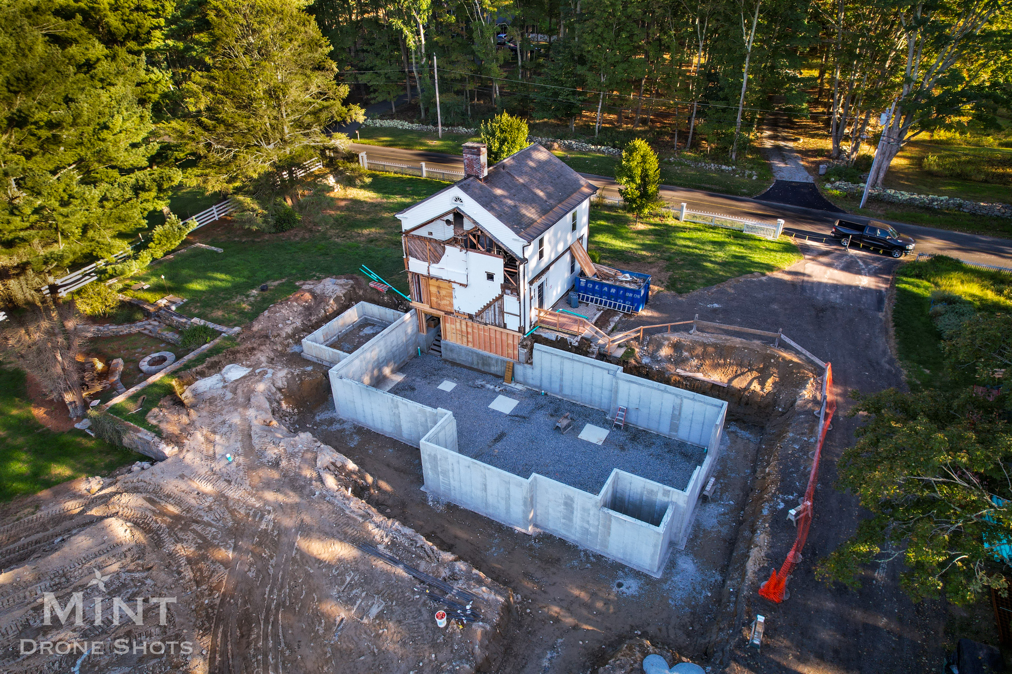 Aerial view of a house under renovation with a large concrete foundation structure in front, surrounded by dirt and construction materials. Trees and a road are in the background.