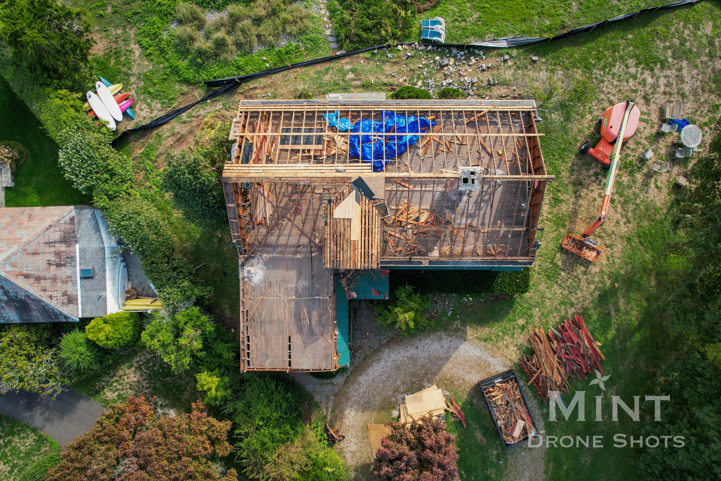 Aerial view of a house under construction with a partially completed roof. Scattered construction materials, wood planks, and machinery are visible. Surrounding greenery includes grass, trees, and shrubs.