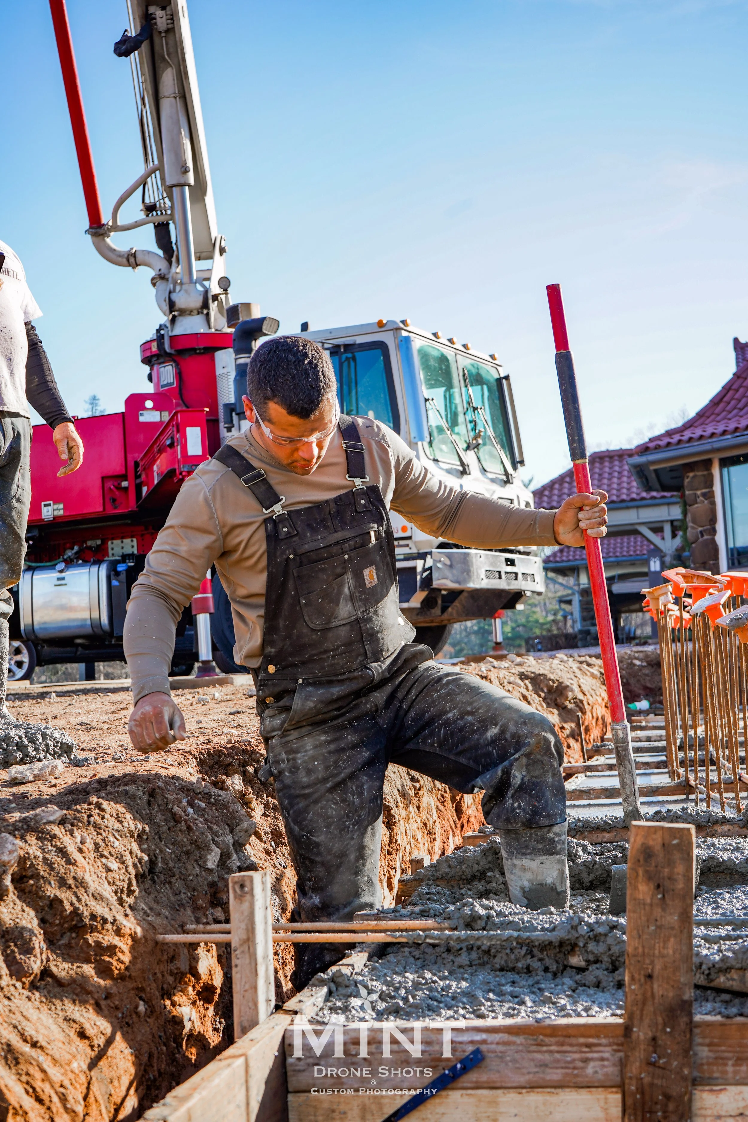 Construction worker wearing overalls and safety goggles standing in wet concrete, with a concrete pump truck in the background, at a construction site under a clear blue sky.