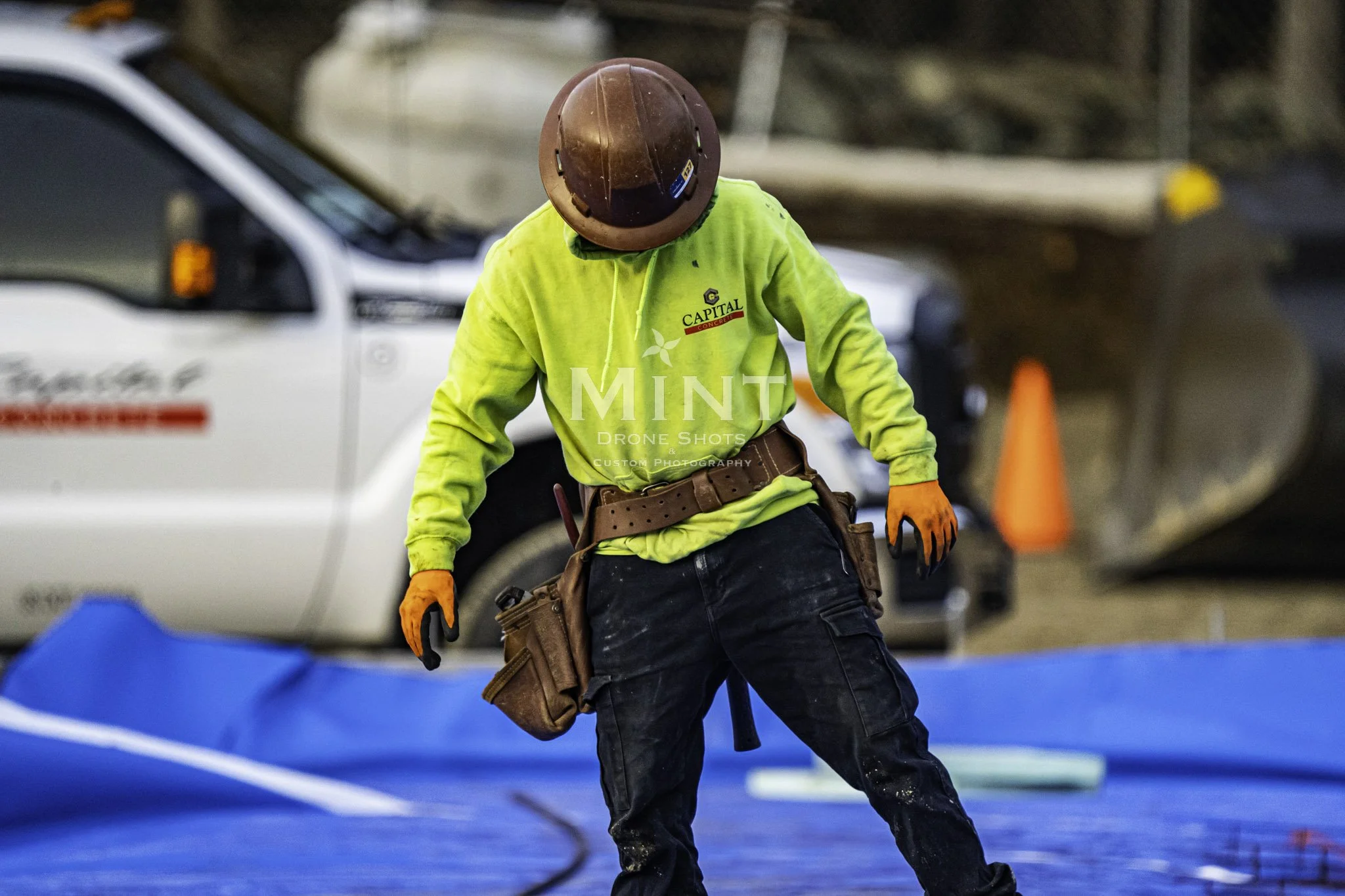 Construction worker in a neon shirt and hard hat working on a site with tools and a utility vehicle in the background.