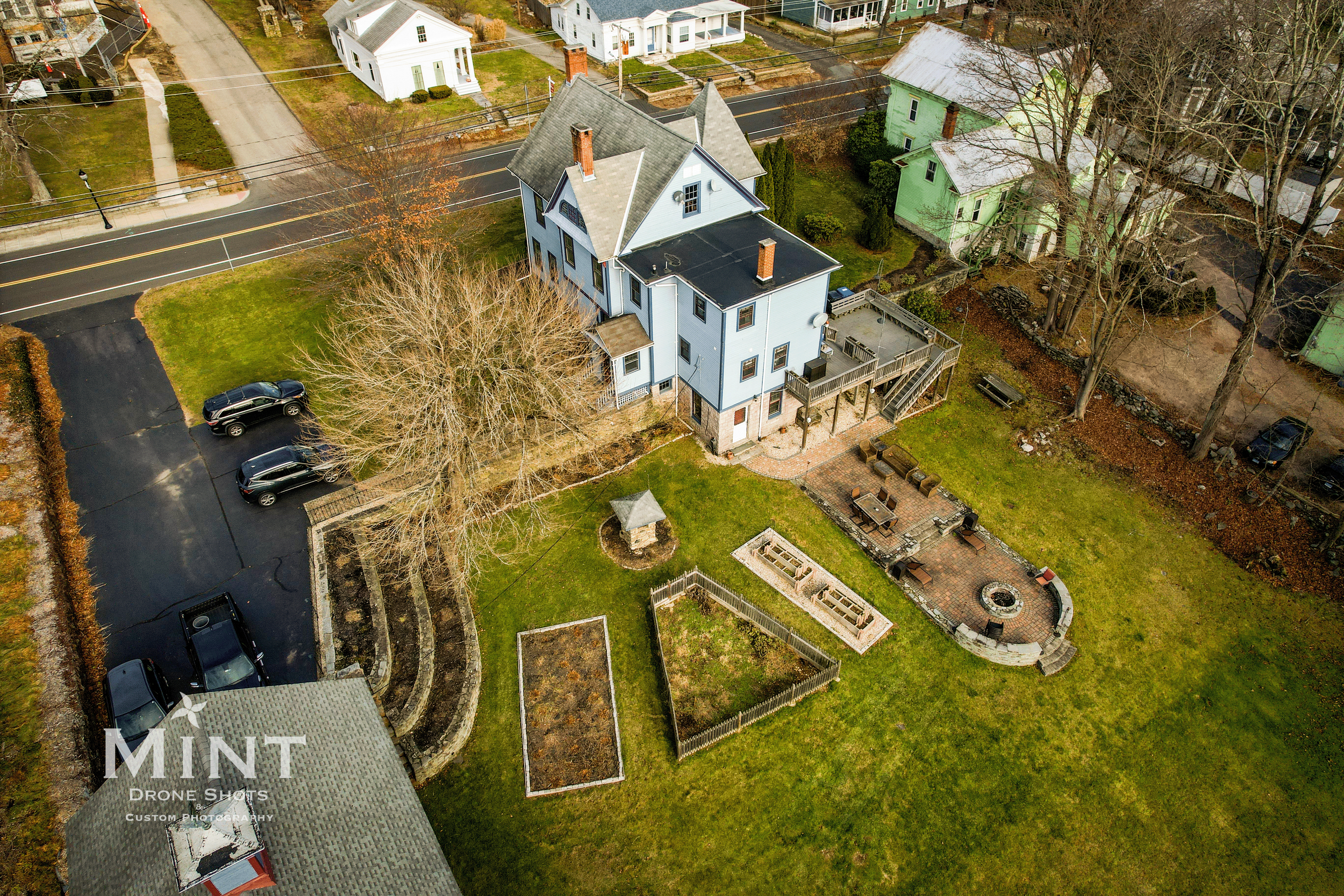 Aerial view of a residential property with a blue multi-story house, landscaped backyard, patio with seating, planters, and trees in a suburban neighborhood.