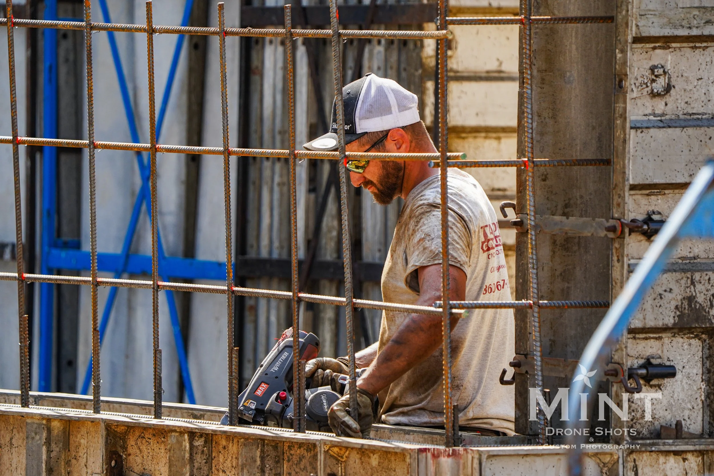 Construction worker using a power tool on a building site, surrounded by rebar and concrete forms.