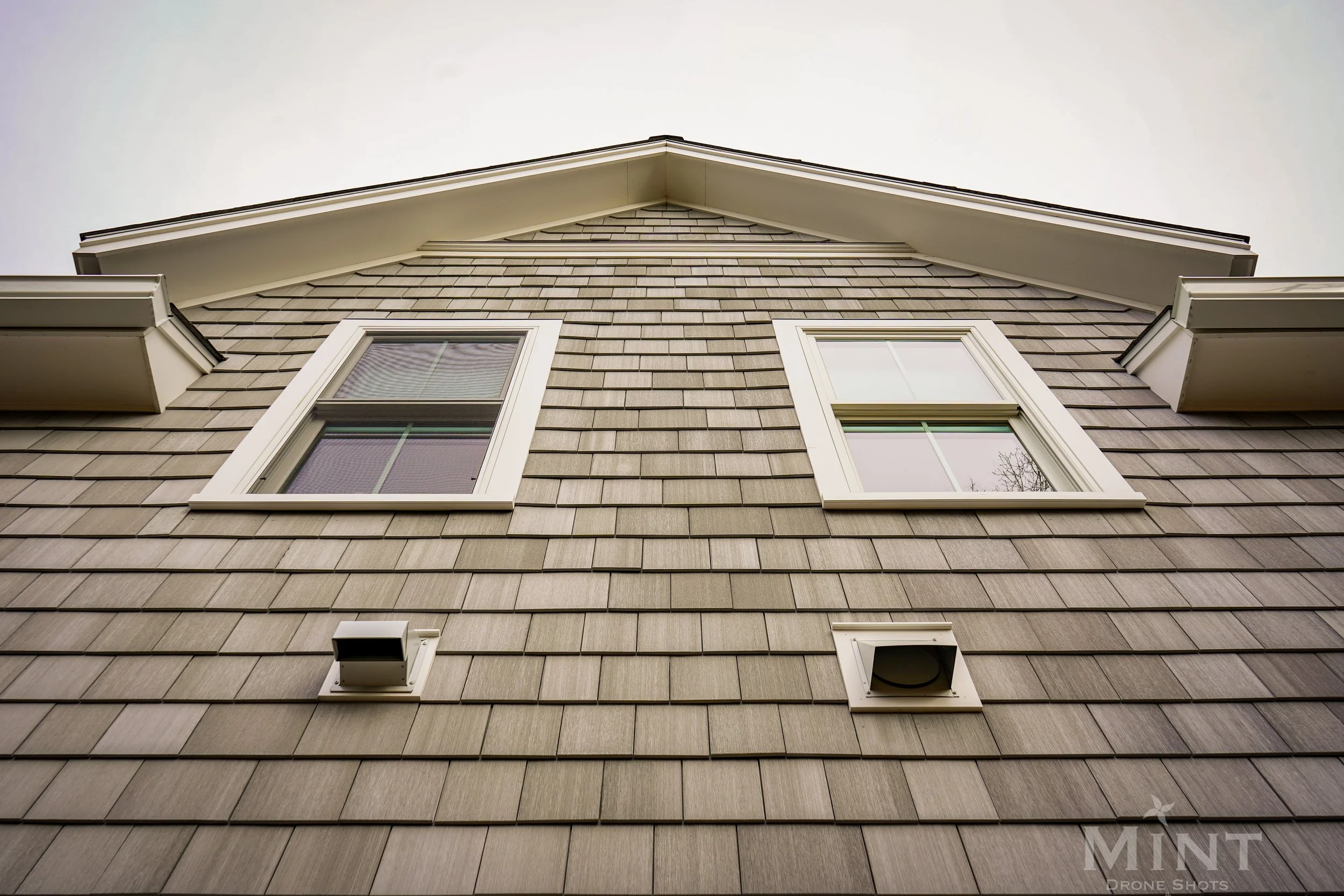 Close-up of a house exterior with shingle siding, two windows, and vents, against a gray sky.