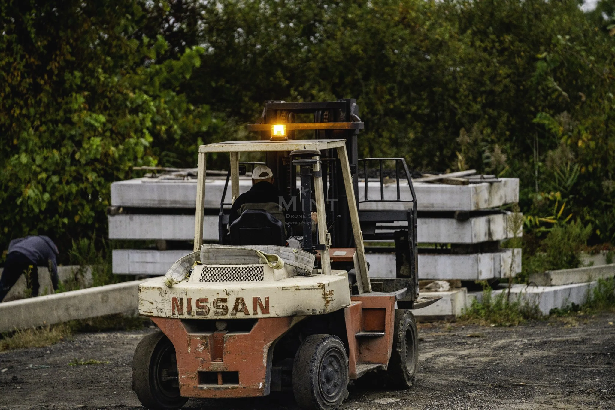 A person operating a Nissan forklift outdoors near stacked concrete blocks, surrounded by greenery.