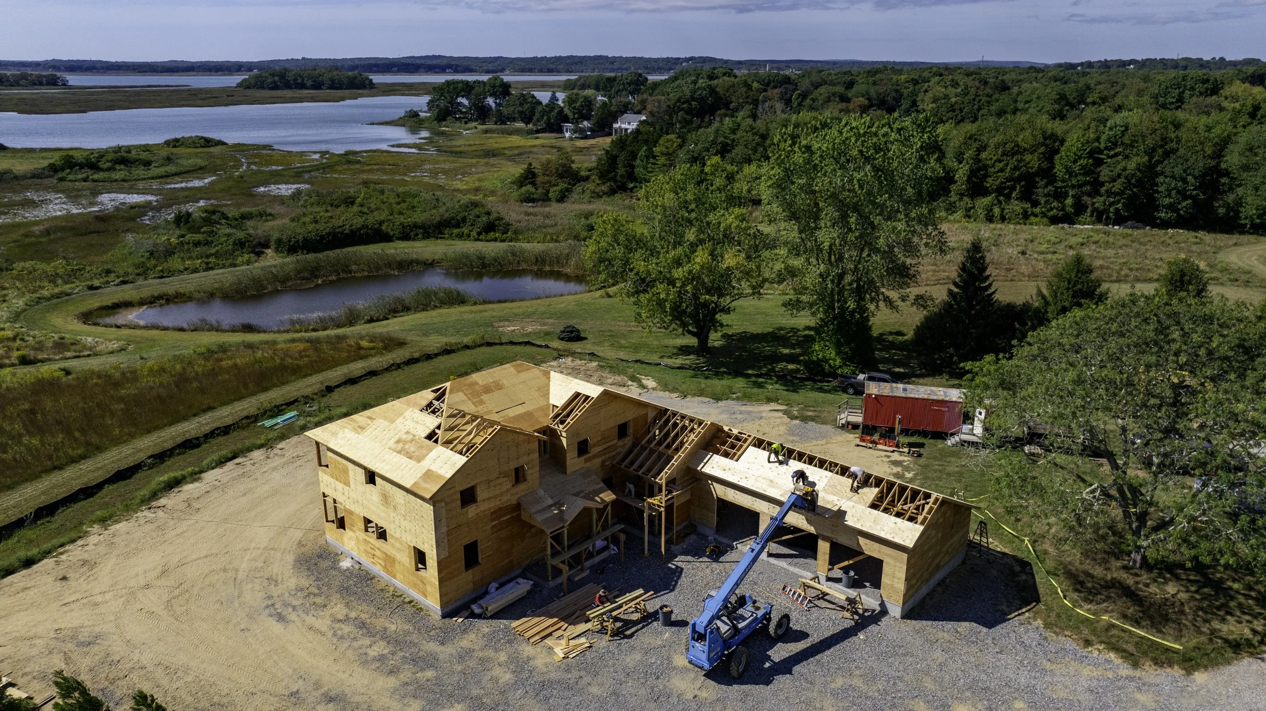 Aerial view of a house under construction with wooden framing, set in a rural landscape near a lake and surrounded by trees and fields.