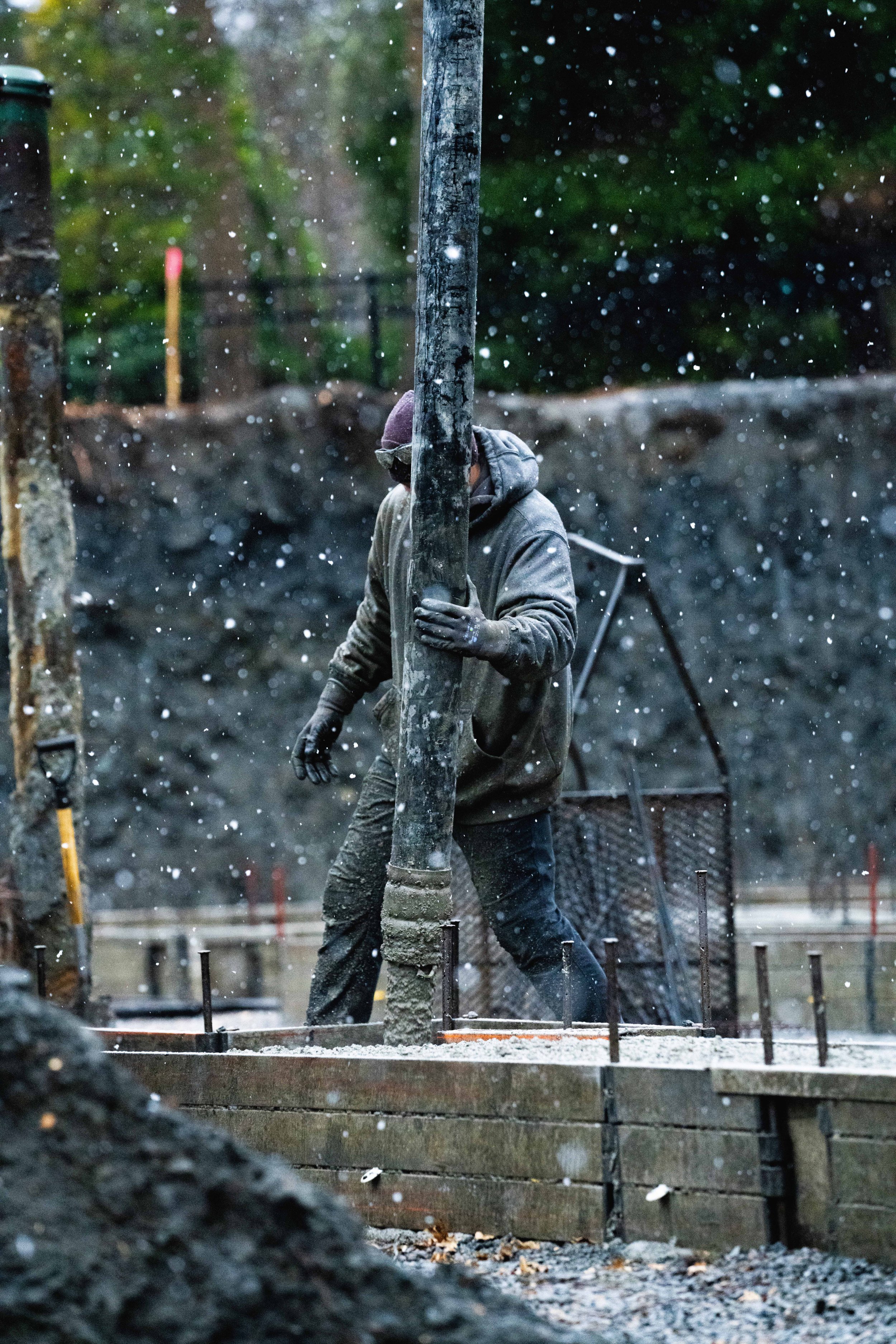 Construction worker wearing a hoodie and gloves, handling a concrete pump during snowy weather.