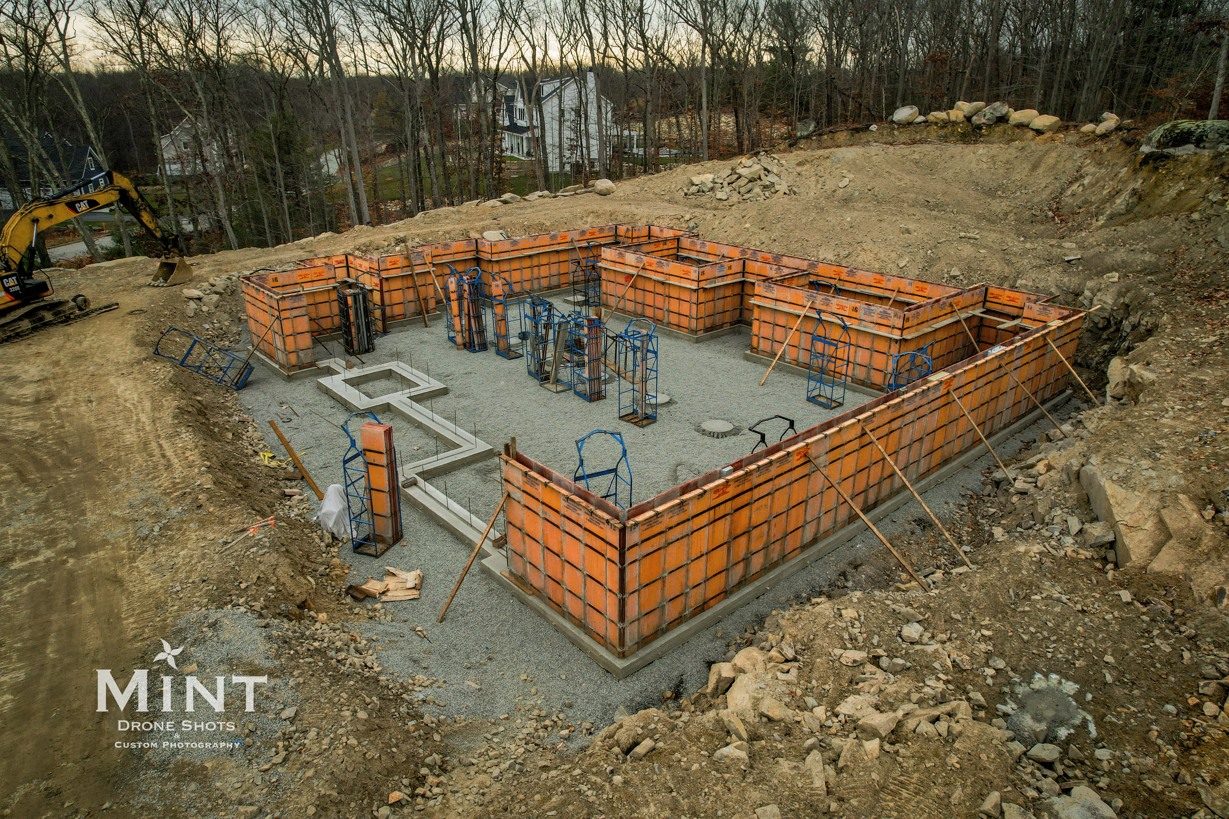 House foundation under construction with orange concrete forms, gravel base, and blue reinforcement bars on a construction site with surrounding trees and a distant house.