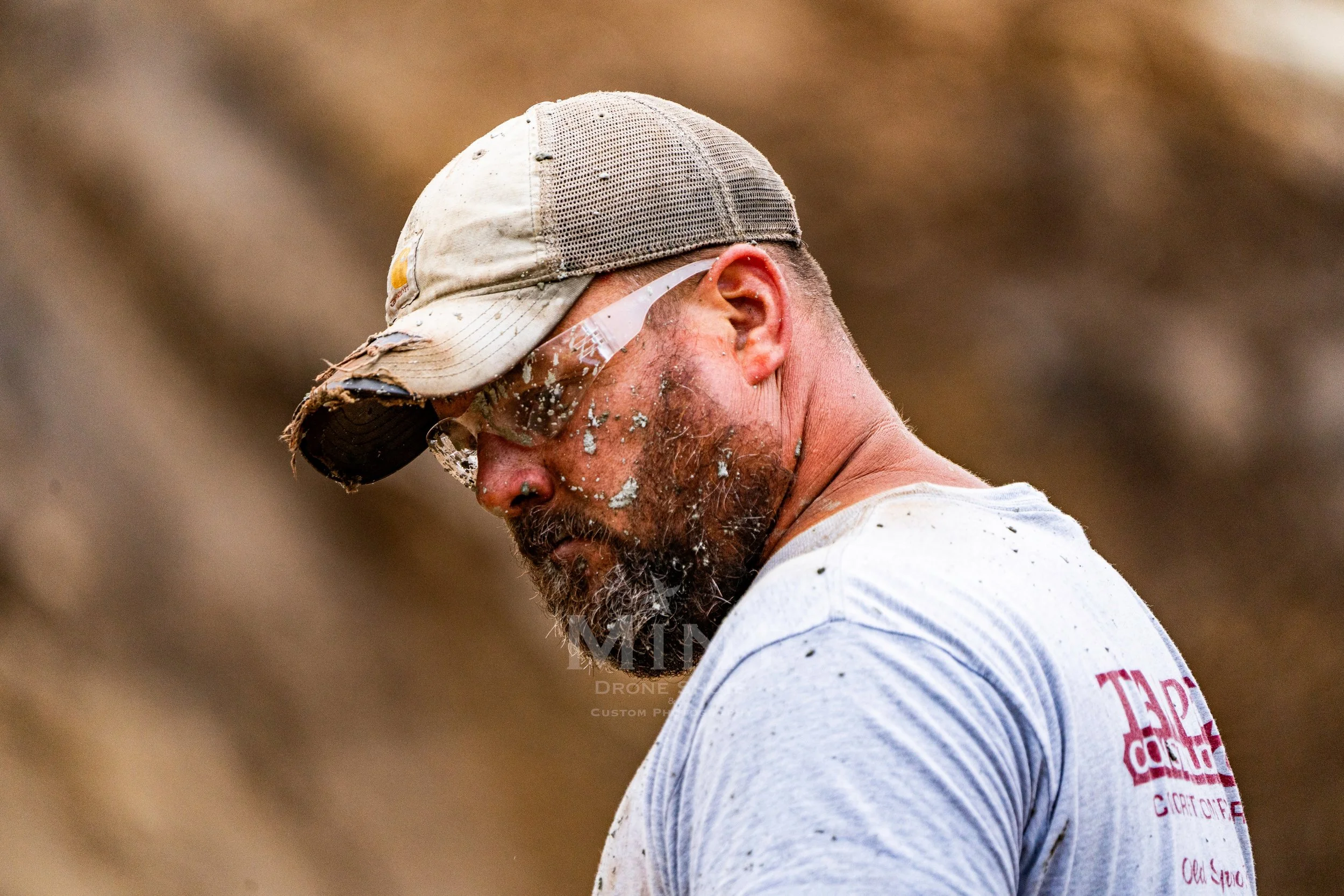 Construction worker with safety glasses and a worn cap, covered in dirt, focusing on work.