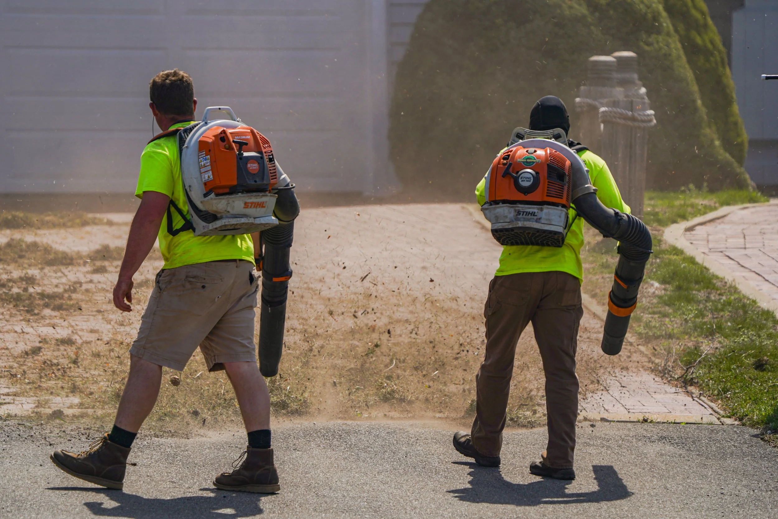 Two workers using backpack leaf blowers to clear debris from a pathway.