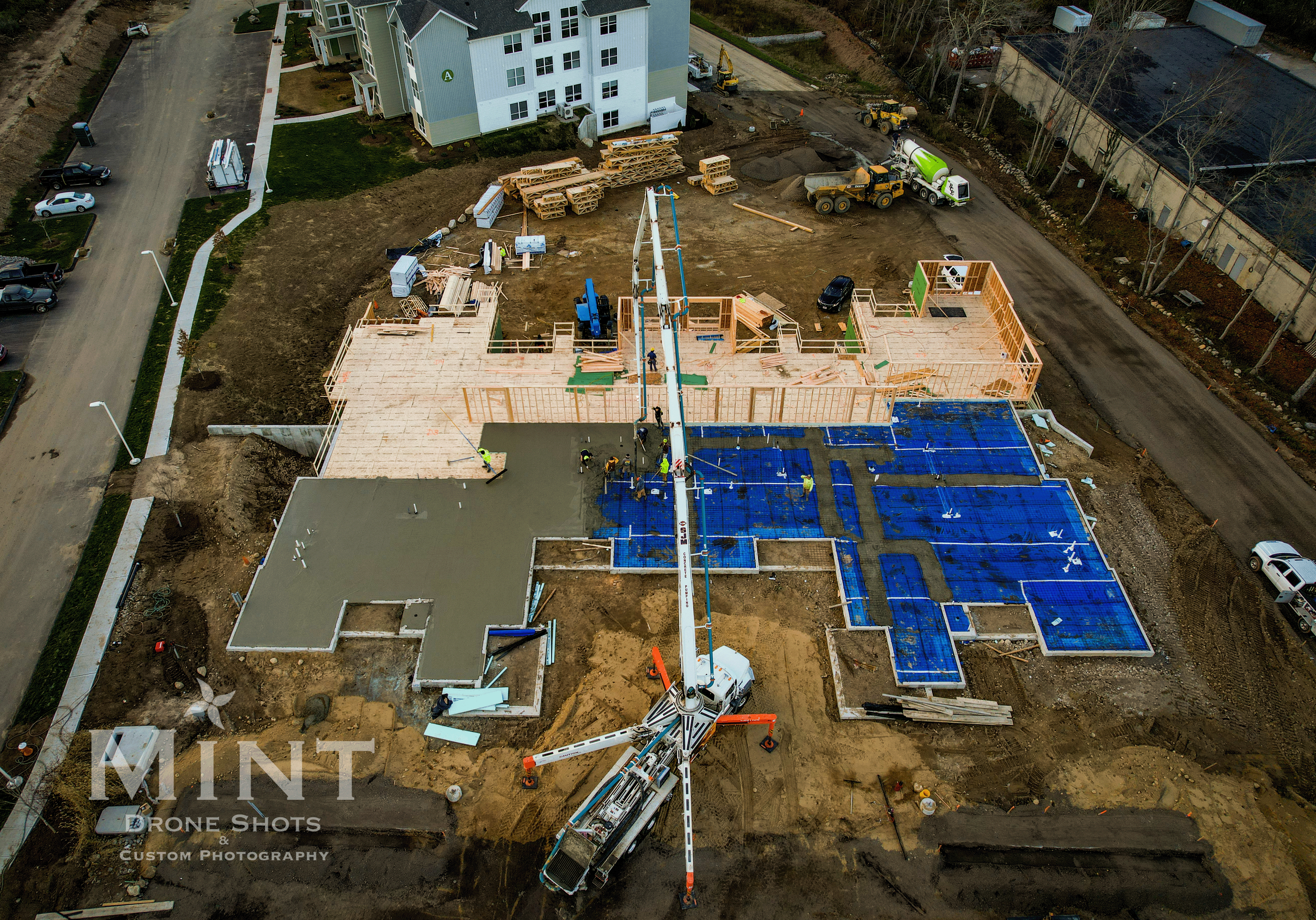 Aerial view of a construction site with partially built structures, framing, concrete slab, and construction equipment. Residential buildings are in the background.