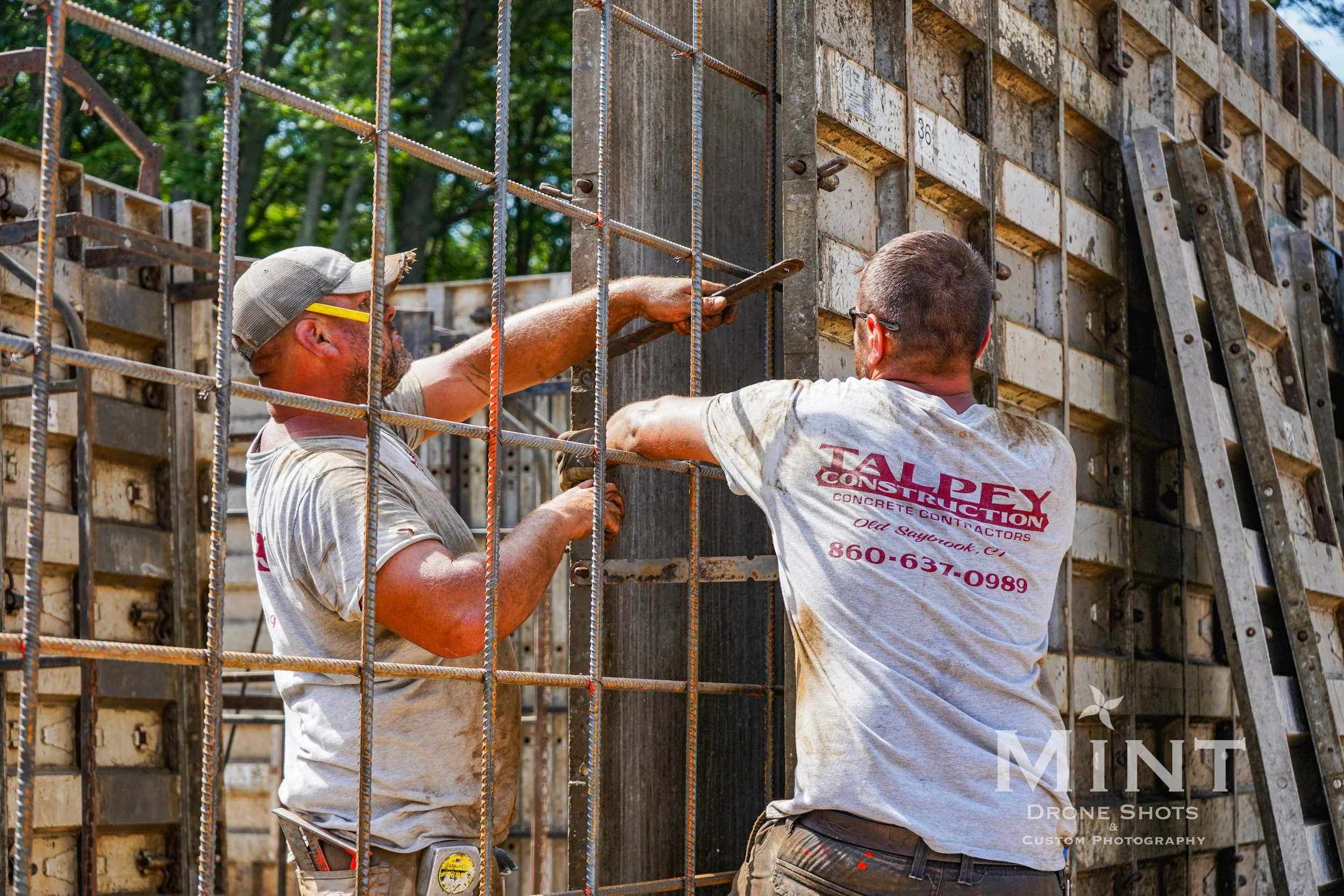 Two construction workers wearing casual work clothes and hats are installing a metal framework at a construction site. The framework is likely for concrete reinforcement. They are engaged in manual labor under clear daylight, with trees visible in th