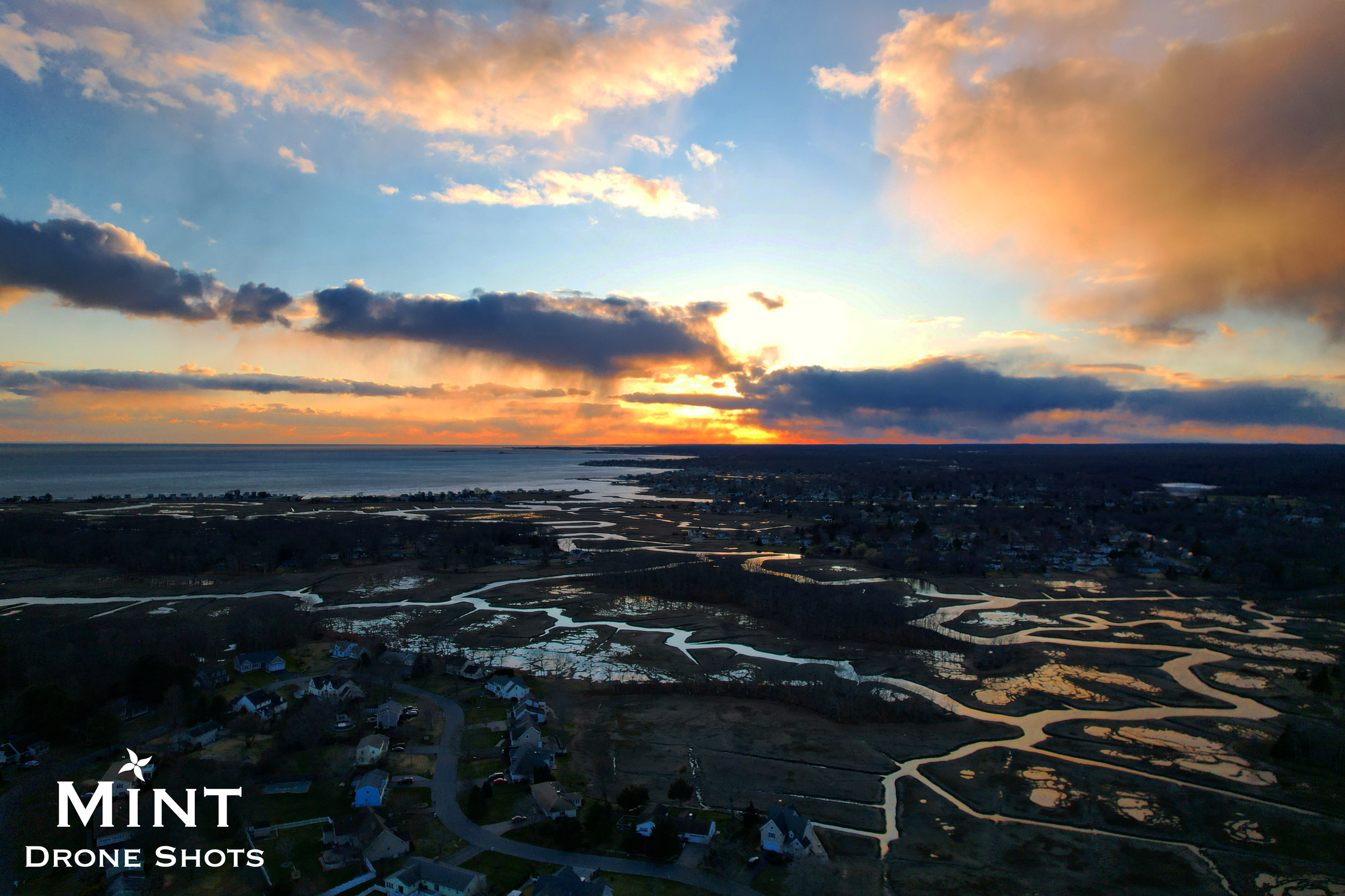 Aerial view of a coastal landscape at sunset with marshlands, winding waterways, and scattered homes. The sky is filled with colorful clouds and the sun setting on the horizon.
