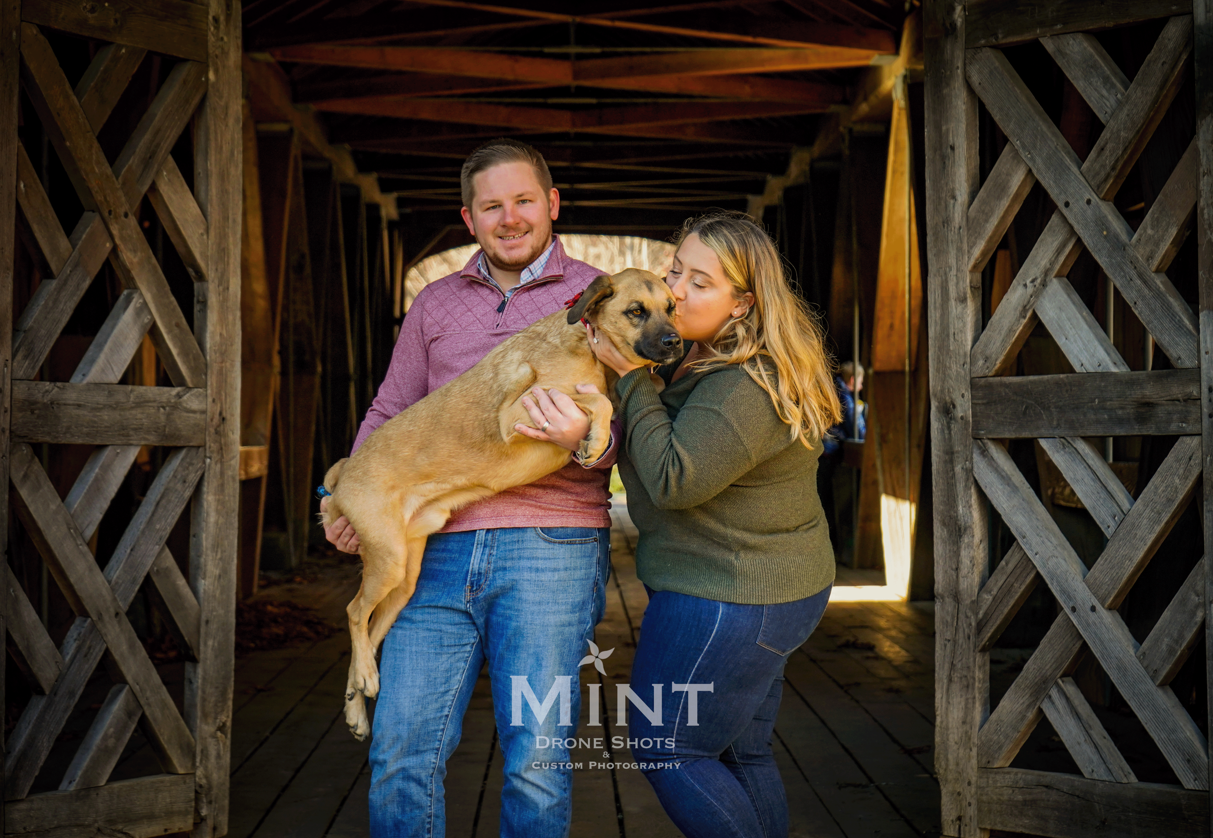 A man and woman holding a large dog in a covered wooden bridge.