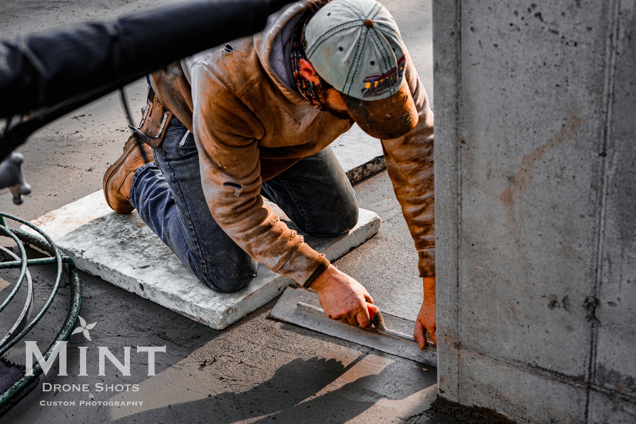 Construction worker smoothing concrete with a trowel on a construction site, wearing a cap and work clothes. Mint Drone Shots watermark visible on image.