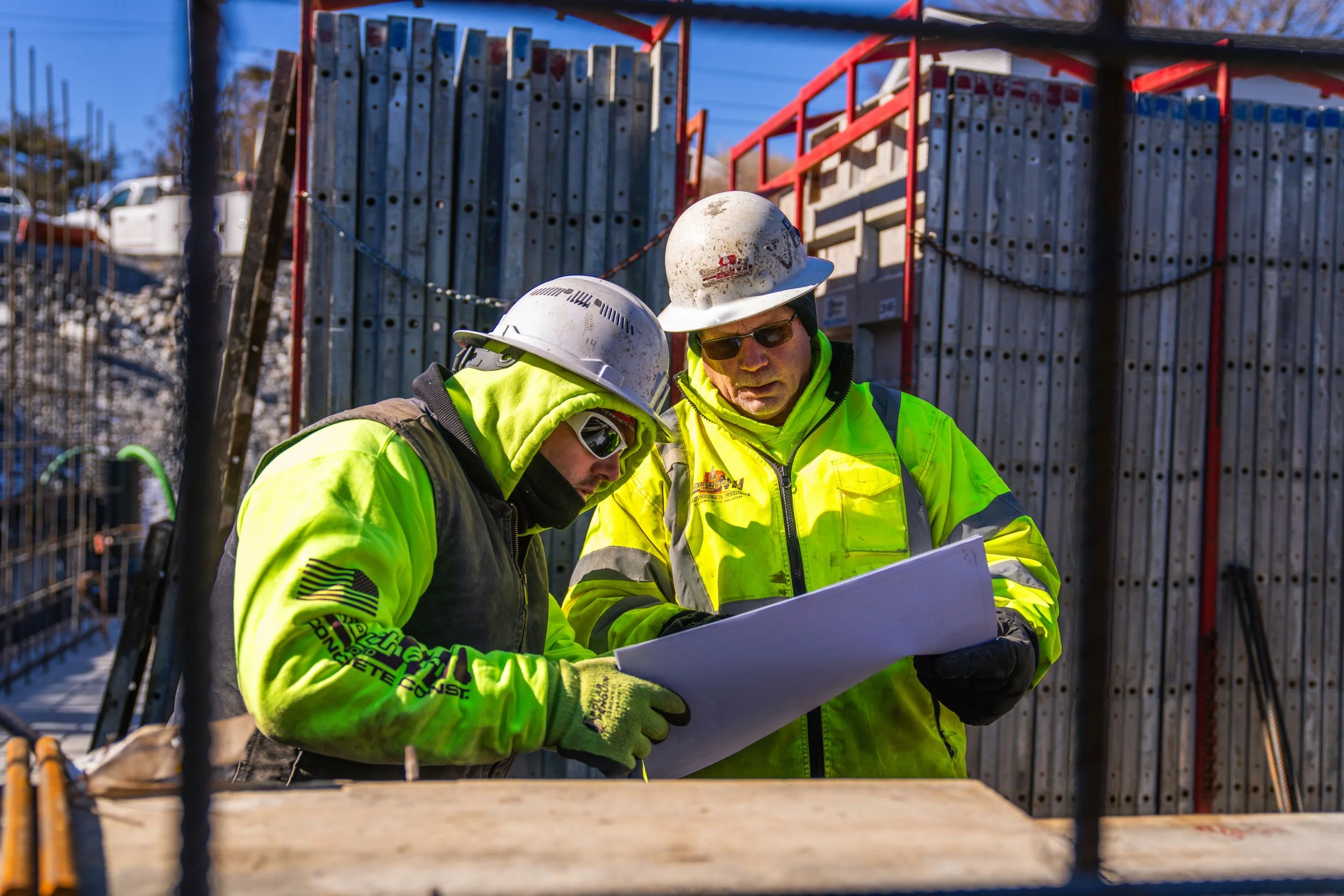 Two construction workers in yellow safety jackets and hard hats reviewing blueprints at a construction site.