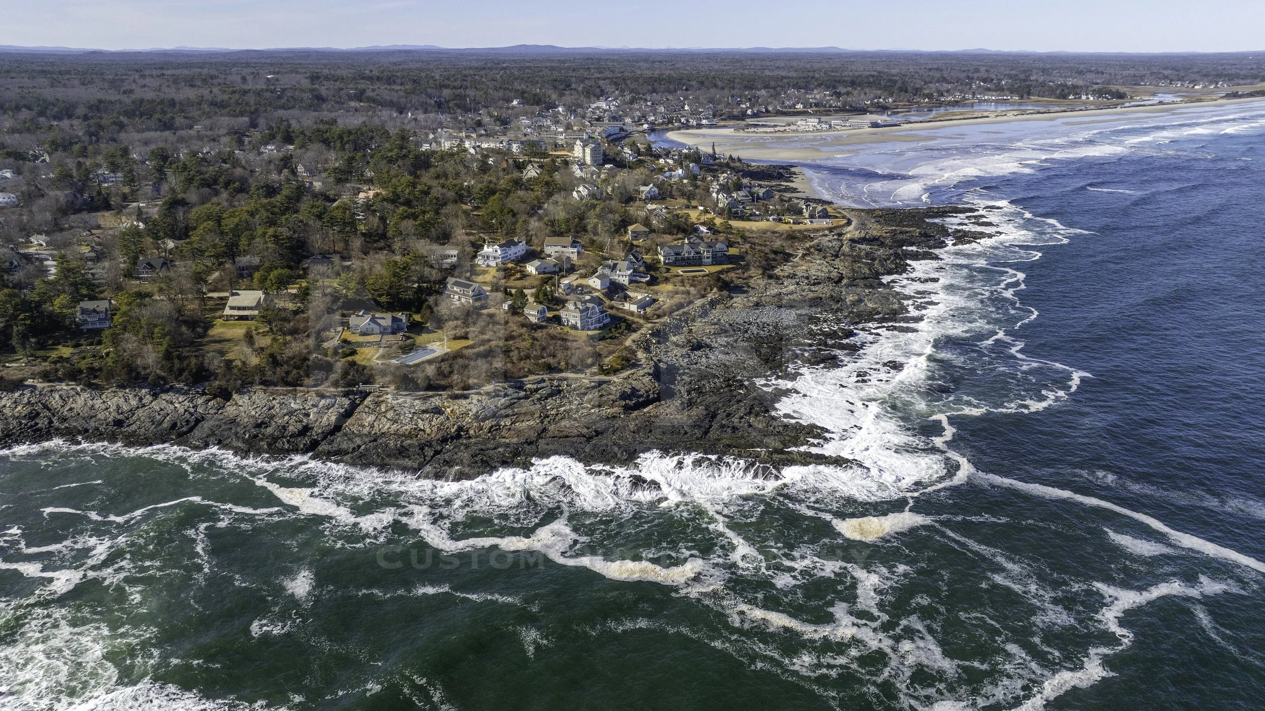 Aerial view of a coastal landscape with ocean waves hitting rocky cliffs and residential houses near the shoreline. The background shows a vast wooded area and a beach along the coast.