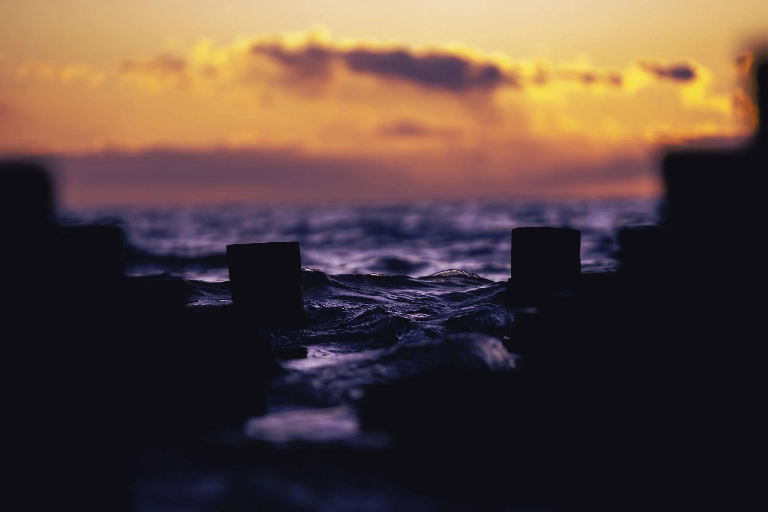 Silhouetted ocean scene at sunset with waves and pilings.