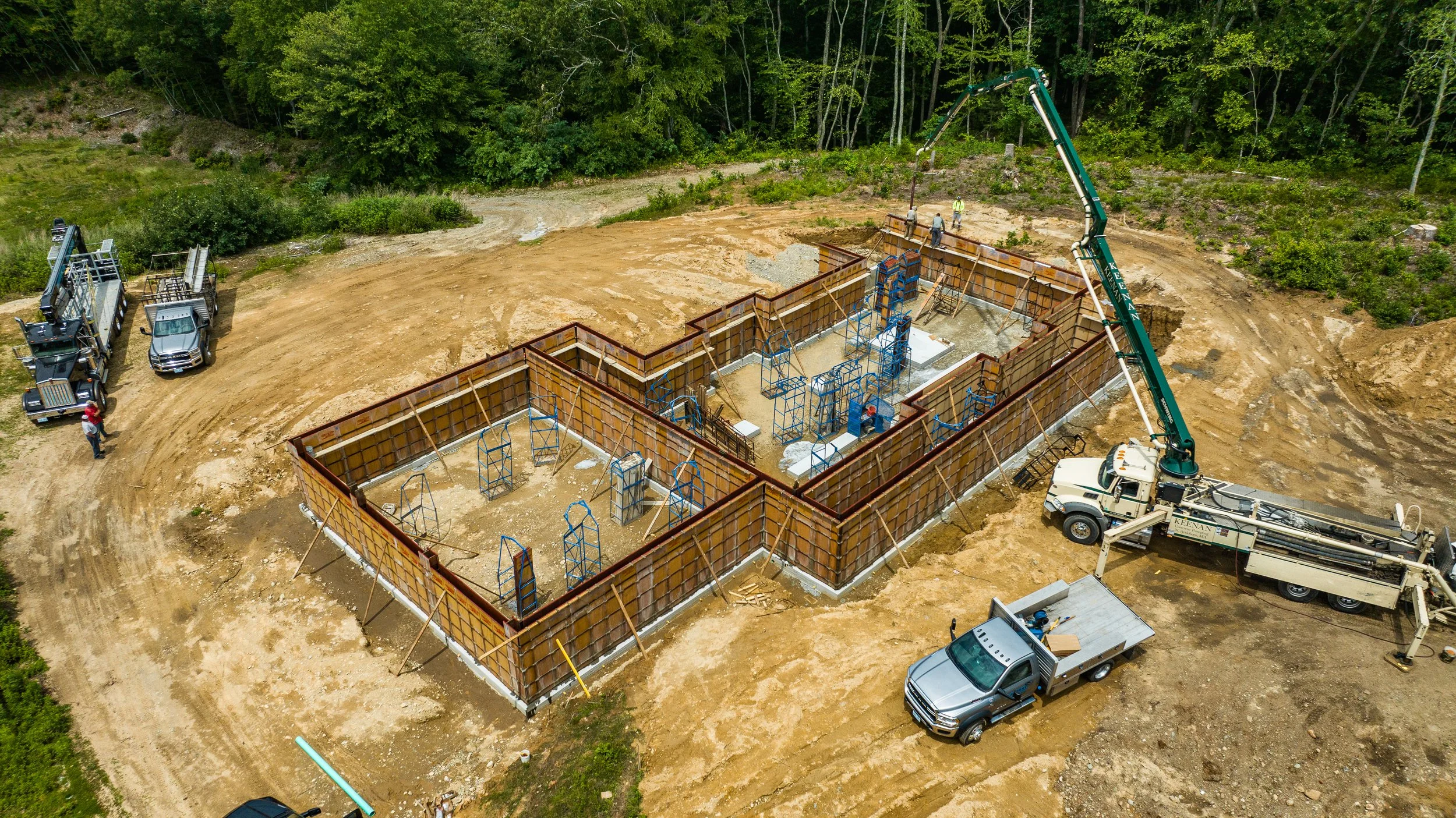 Aerial view of a construction site with concrete foundation forms in place, vehicles and machinery on-site, surrounded by wooded area.