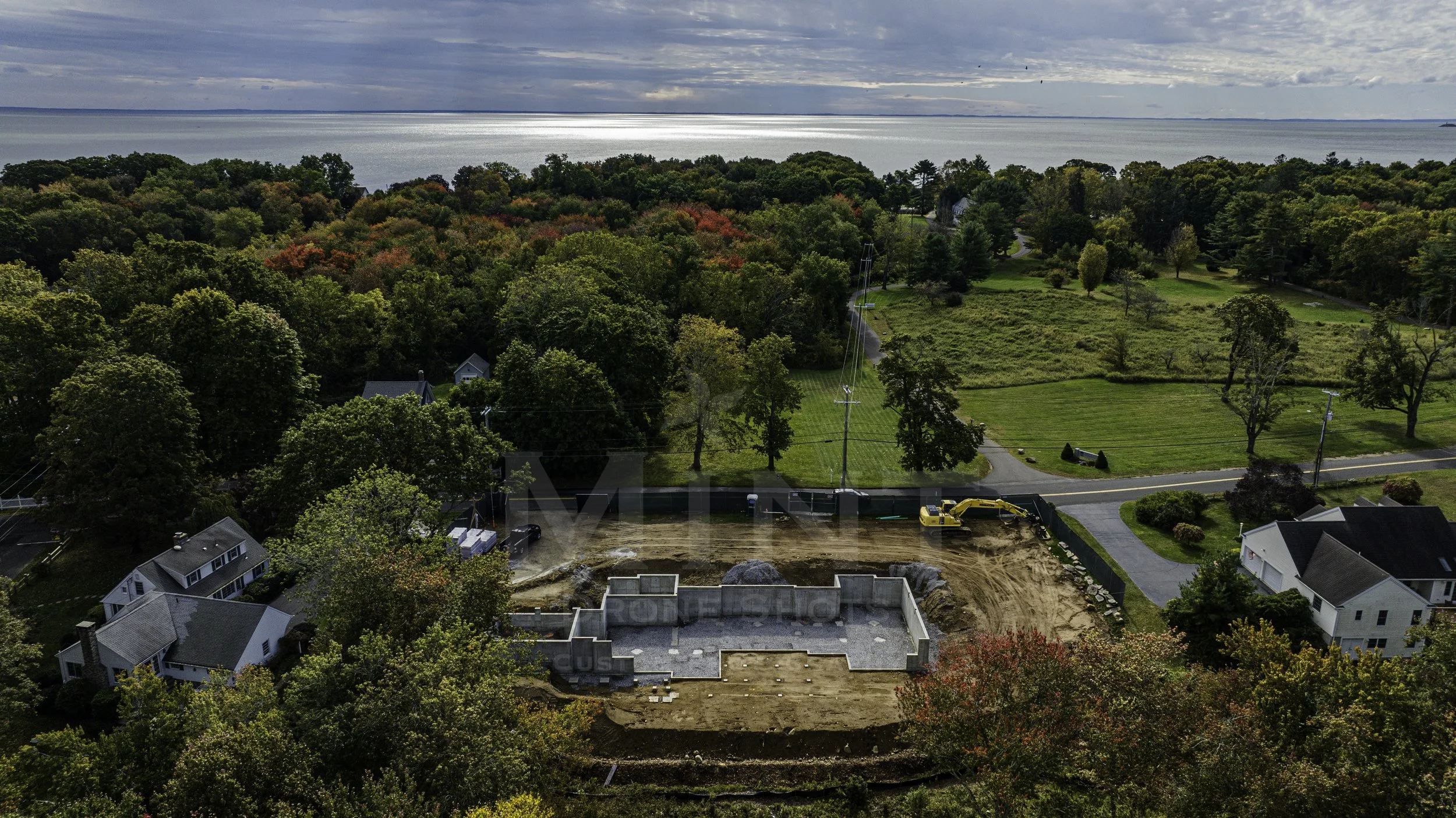 Aerial view of a construction site near a residential area with trees and a road. A foundation is visible, surrounded by dirt. There are houses nearby and a body of water in the distance.