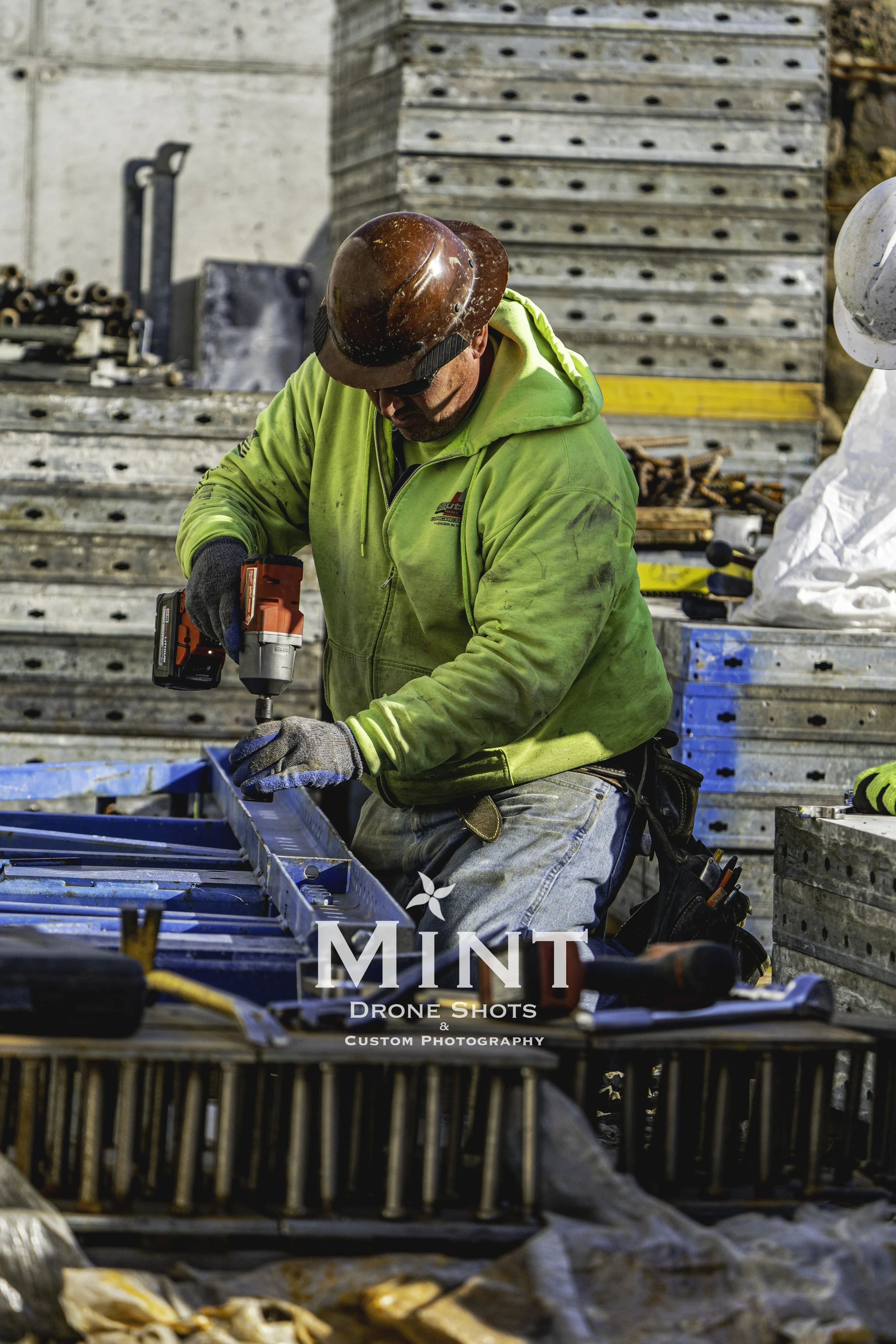 Construction worker wearing a hard hat and reflective jacket using a power drill on metal framework.