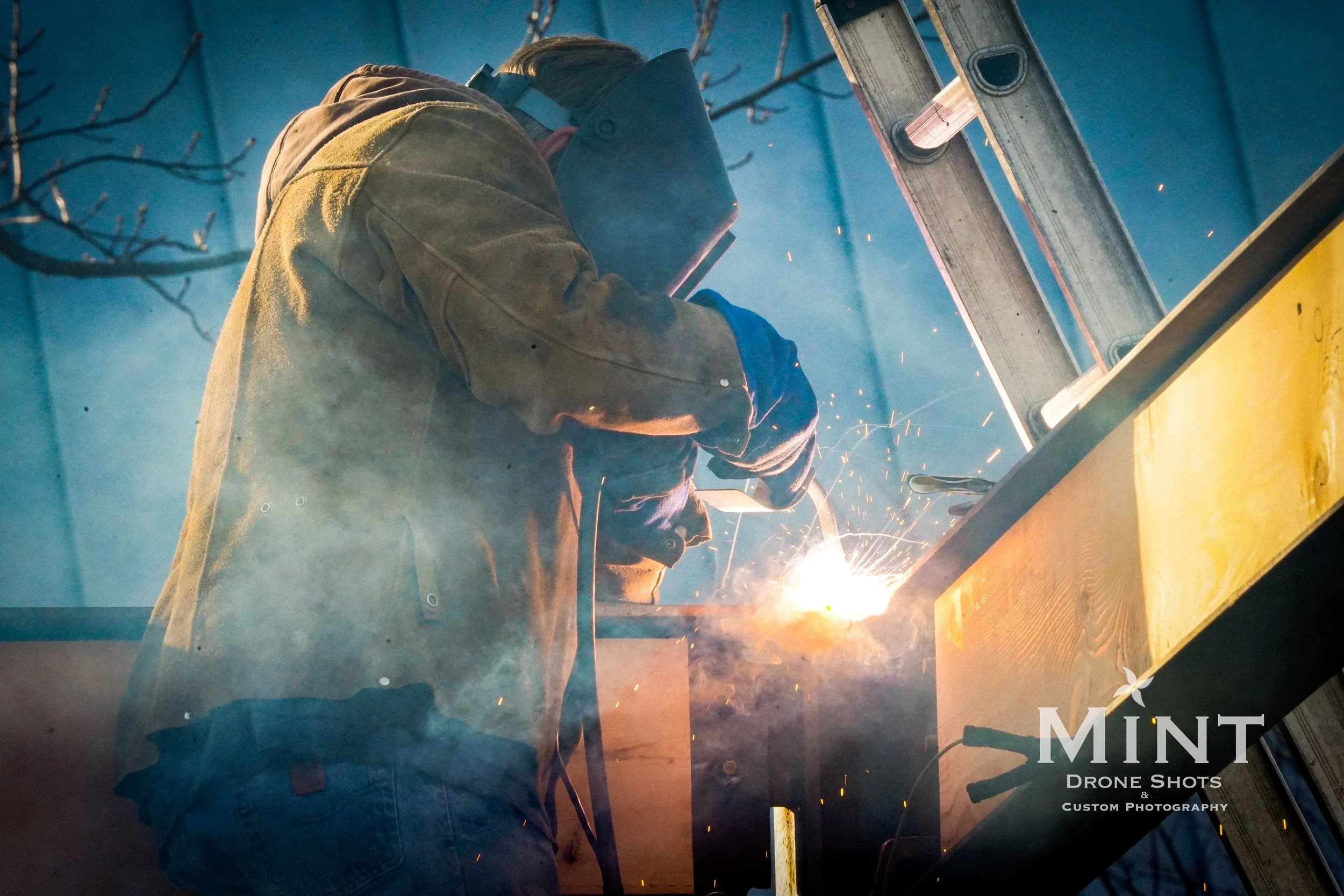 Person wearing welding gear, including helmet and gloves, engaging in welding work outdoors beside a ladder, with sparks and smoke visible.