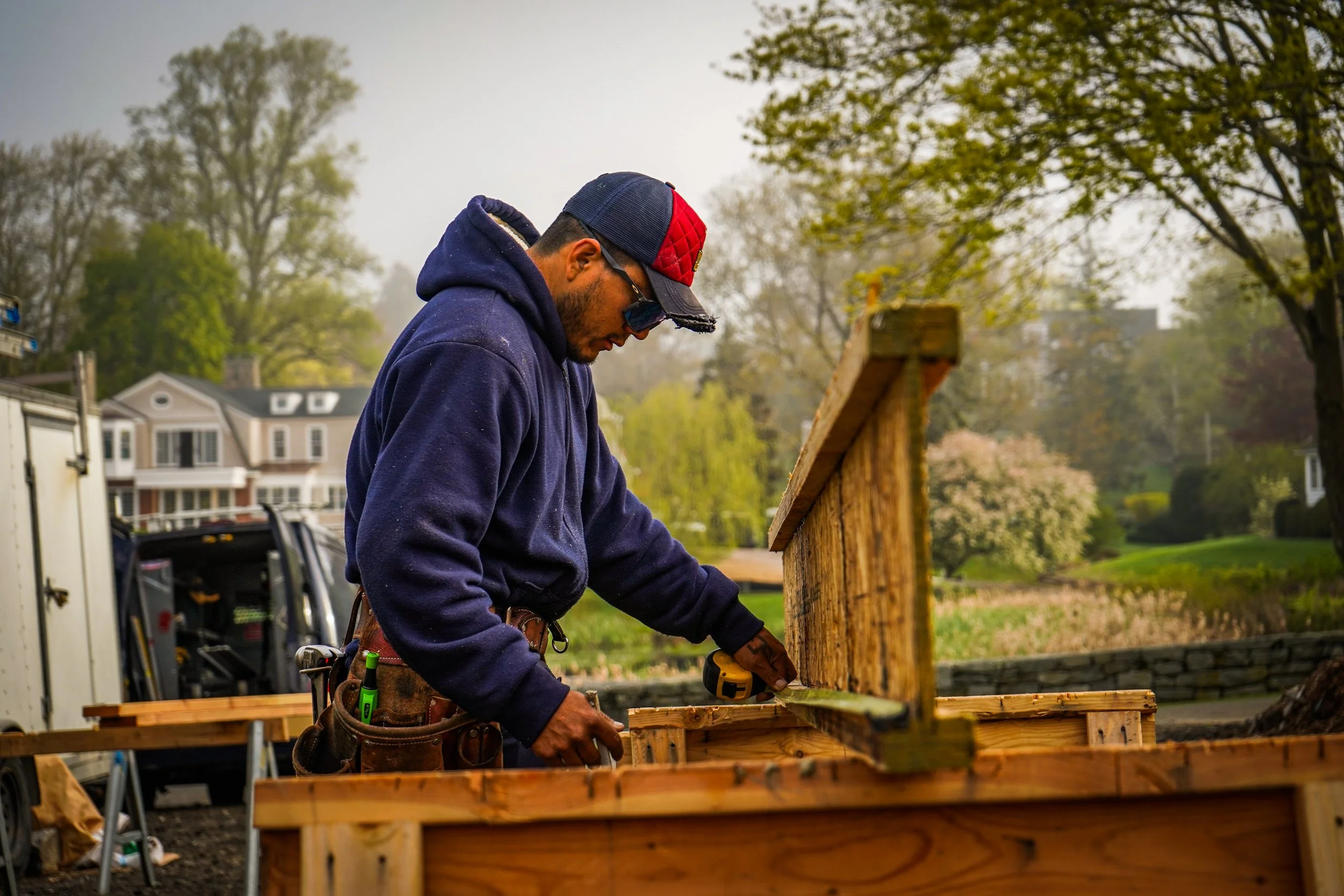 Carpenter working outdoors on wooden structure next to a toolbox, wearing a cap and safety glasses, with trees and houses in the background.